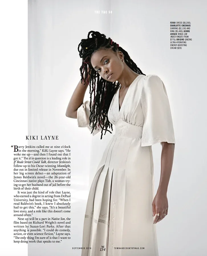 A woman with long dreadlocks wearing a white dress with puffed sleeves, posing with her hand on her head.