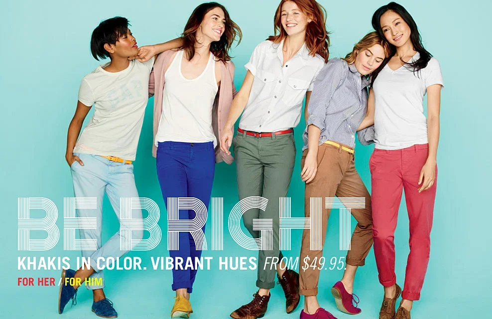 Group of five women wearing colorful khakis standing against a teal background, smiling, with text promoting vibrant-colored khakis for sale.