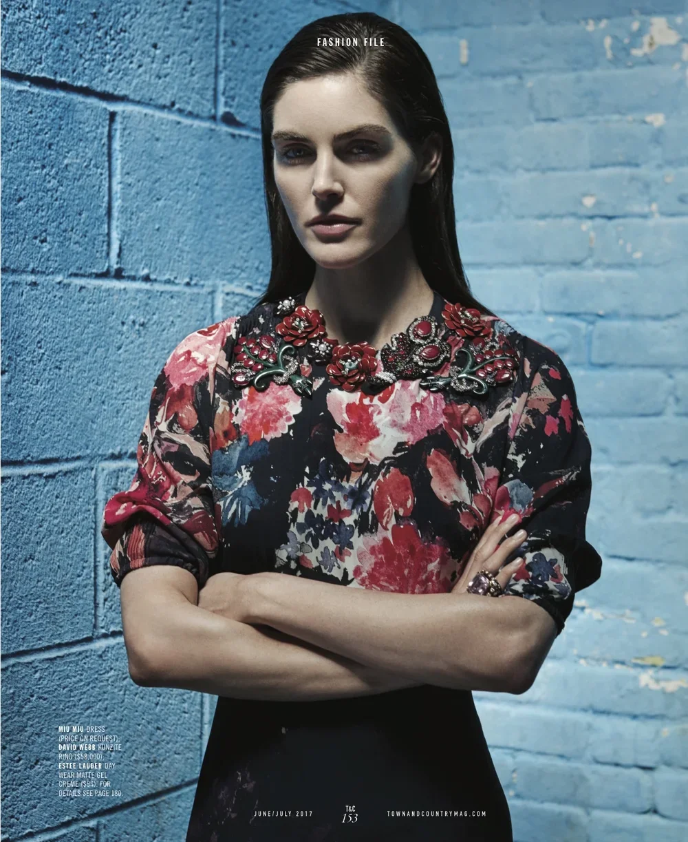 A woman with dark hair and a floral top standing against a textured blue brick wall. She has a confident expression with crossed arms, wearing ornate jewelry on her neck and fingers.