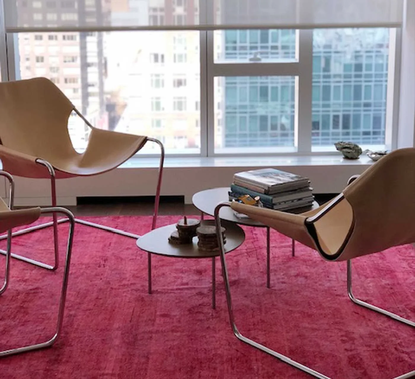 Modern living room with two wooden lounge chairs, a small round coffee table with stacks of magazines and a ceramic ashtray, large windows showing a cityscape, and a pink rug.
