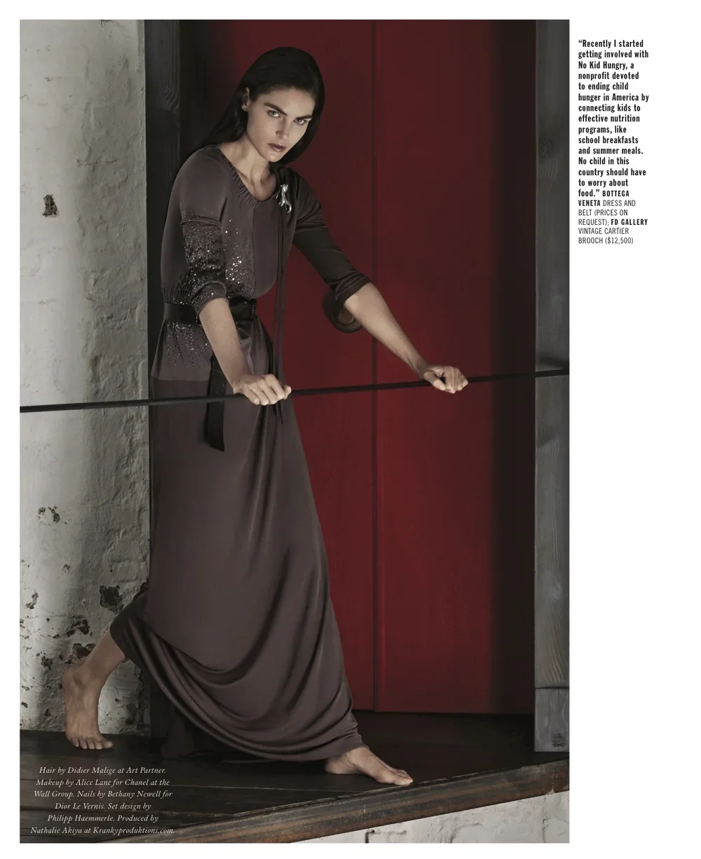 A woman with dark hair, wearing a long, brown dress with sequin details on the sleeves and waist, stands barefoot on a wooden surface, holding a rope with both hands. She is positioned against a textured concrete wall and a red accent wall, looking d