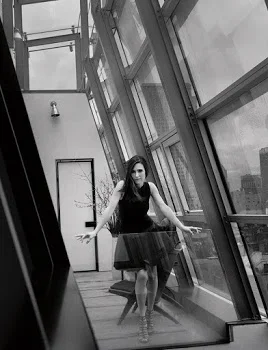 A woman in a black dress standing in a modern glass corridor with large windows and metal framing.