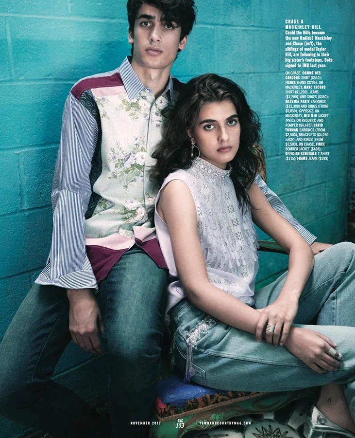 A young man and woman sitting against a teal brick wall. The man has dark curly hair and wears a colorful jacket with floral and striped patterns and jeans. The woman has wavy dark hair, big earrings, a white lace top, denim shorts, and multiple ring