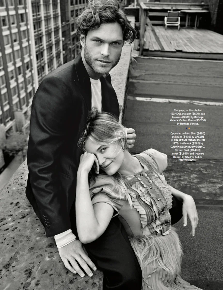 Black and white photo of a man with tousled hair and light beard sitting on a rooftop. He is wearing a jacket over a shirt. A young girl with long, light hair and a dress sits on his lap, leaning her head against his shoulder while smiling and winkin