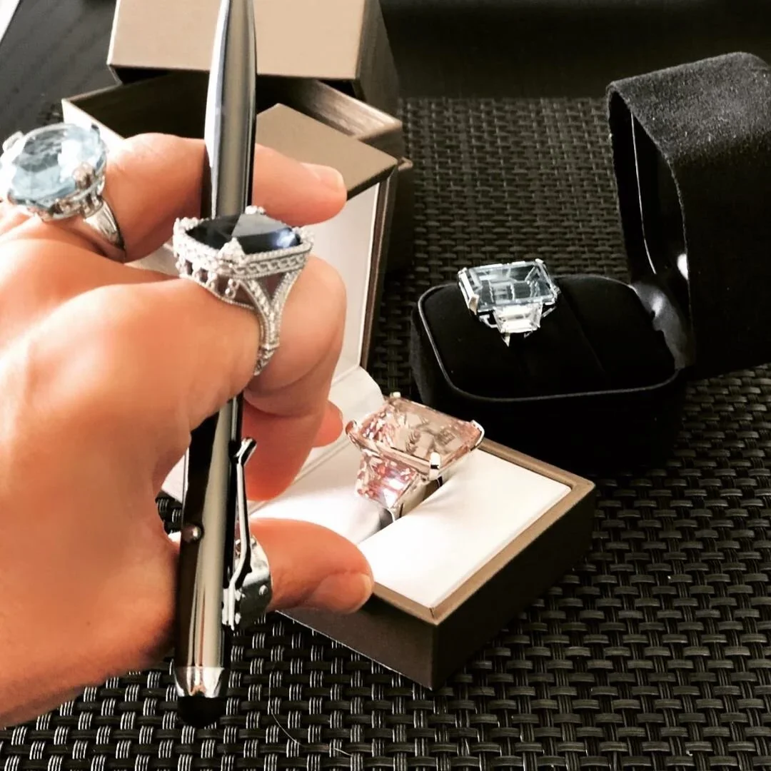Hand holding a silver pen and wearing large statement rings, including one with a large black gemstone, above an open jewelry box with rings, including a large black gemstone ring, placed on a black woven surface.