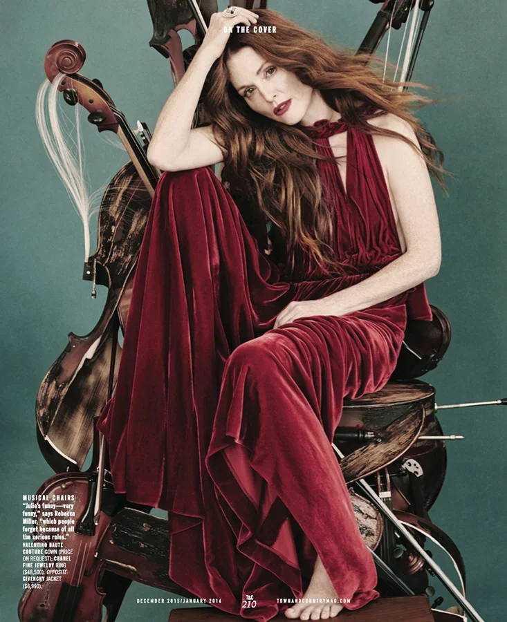 Woman with long red hair in a red velvet dress sitting on a wooden chair surrounded by orchestral string instruments, posing with her hand on her head.