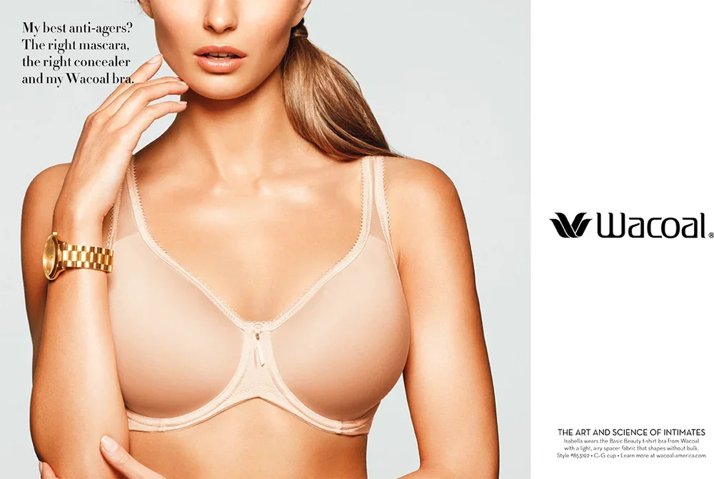 A woman wearing a nude-colored Wacoal underwire bra, with a gold watch on her left wrist, touching her face with her left hand, and the Wacoal logo visible on the right side.