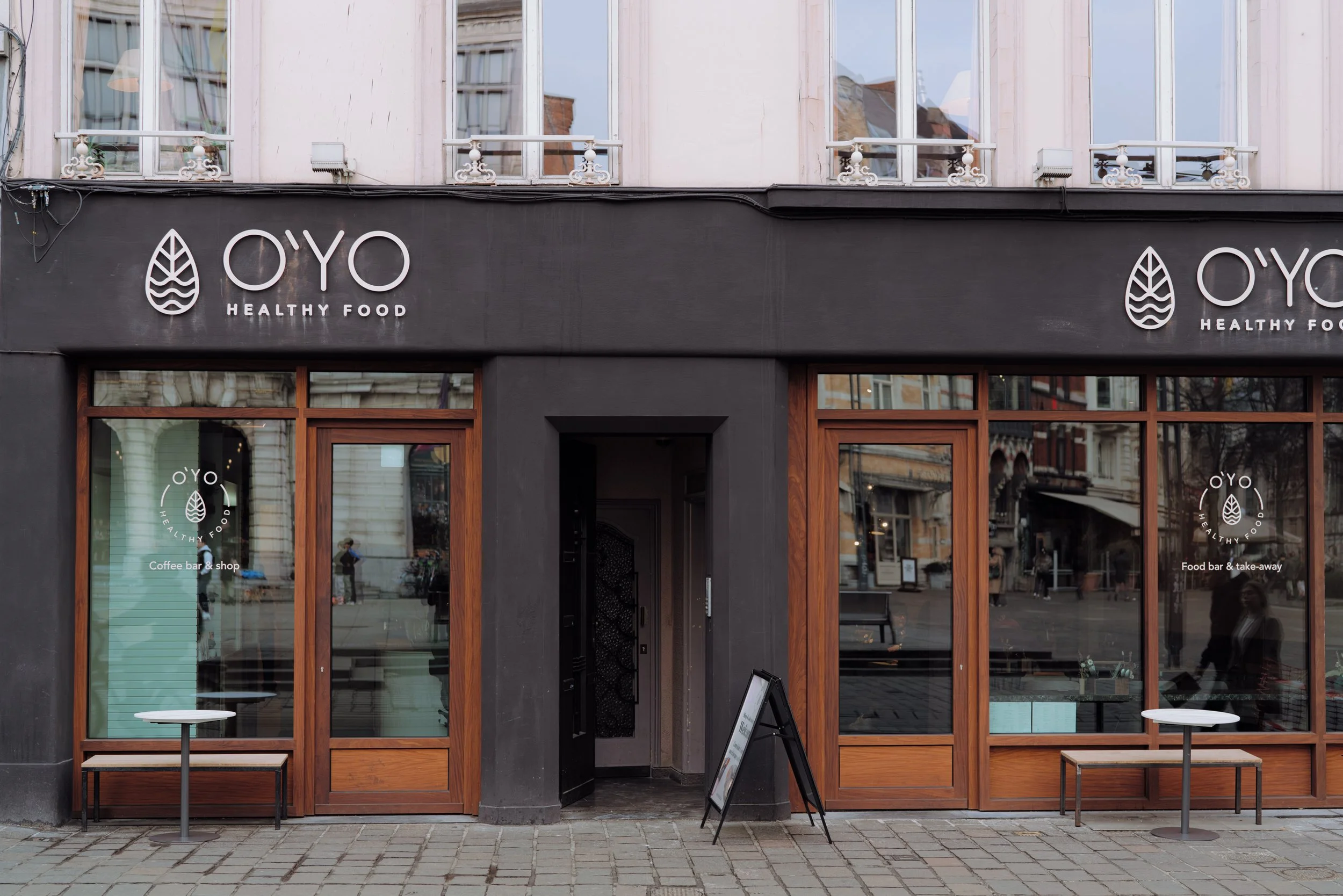 The facade of a black cafe with large wooden framed glass windows and a door, labeled 'OYO Healthy Food', with outdoor tables on a paved sidewalk.