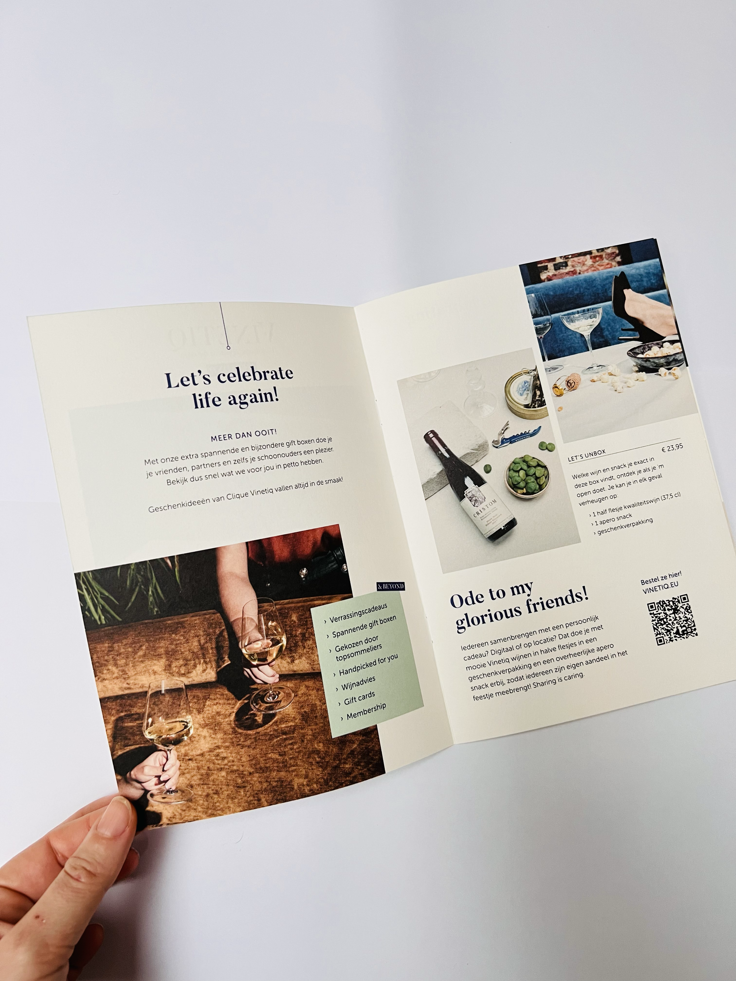 Open brochure with images of wine bottles, glasses, and snacks, text in Dutch and English celebrating life, friends, and gifting ideas, featuring a picture of two hands holding glasses of white wine against a wooden table.