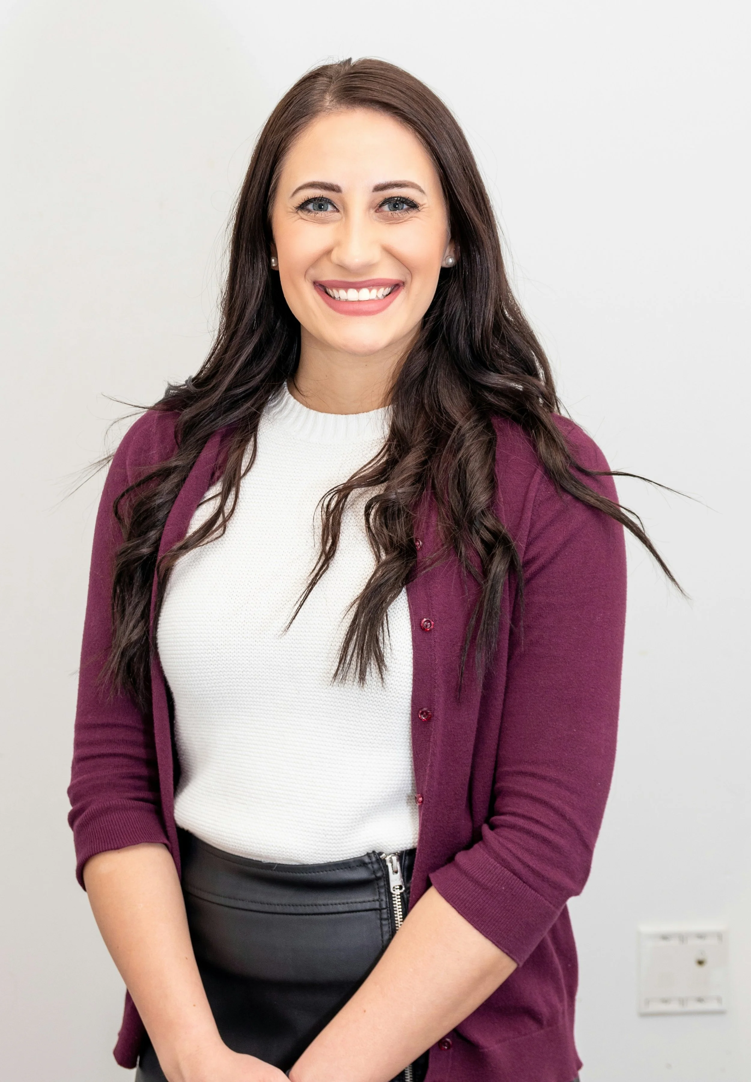 A young woman with long dark brown hair, smiling, wearing a white top, maroon cardigan, and black pants, standing against a plain white wall.