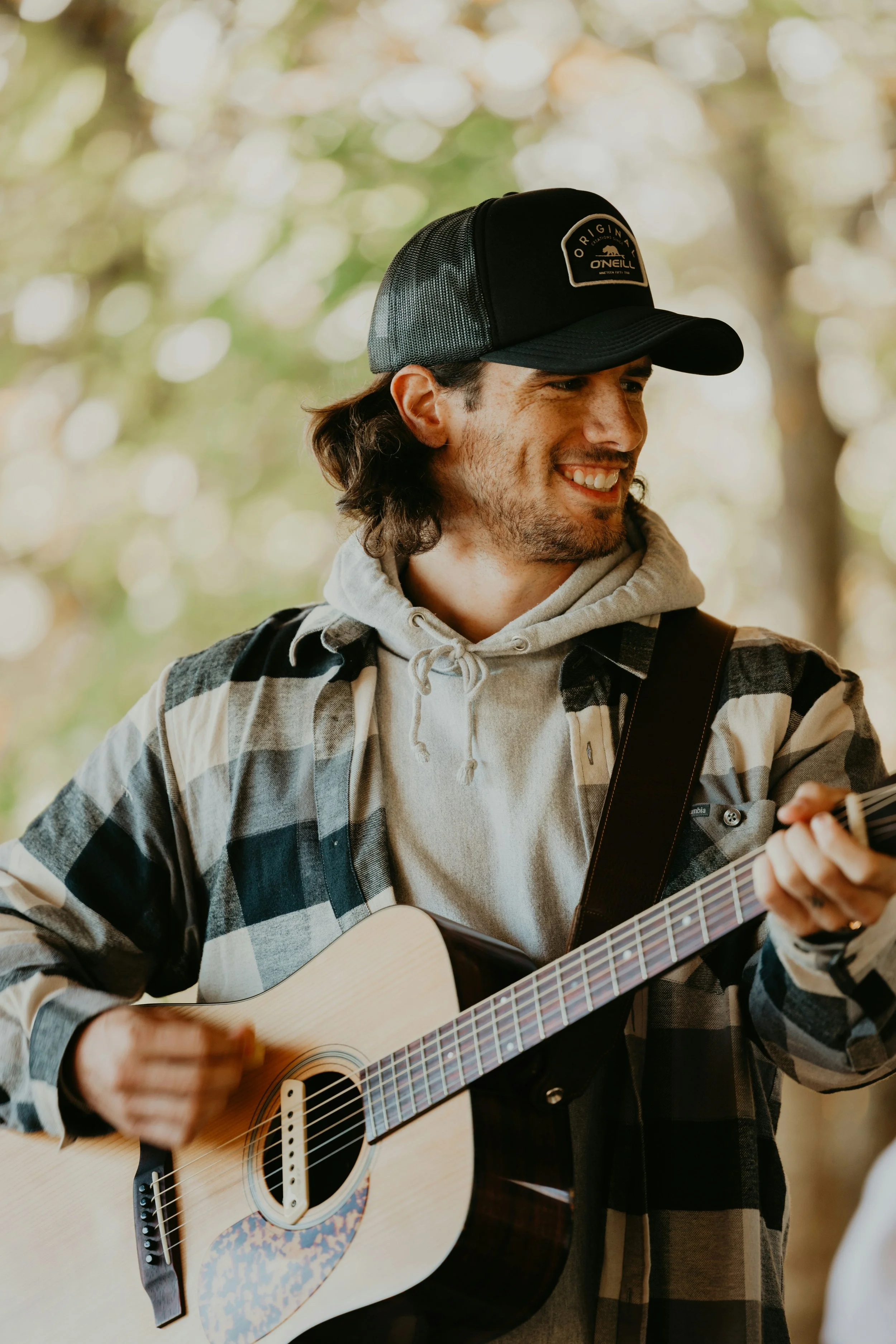 A young man wearing a black trucker hat, a gray hoodie, and a black and white flannel shirt playing an acoustic guitar outdoors in a wooded area.