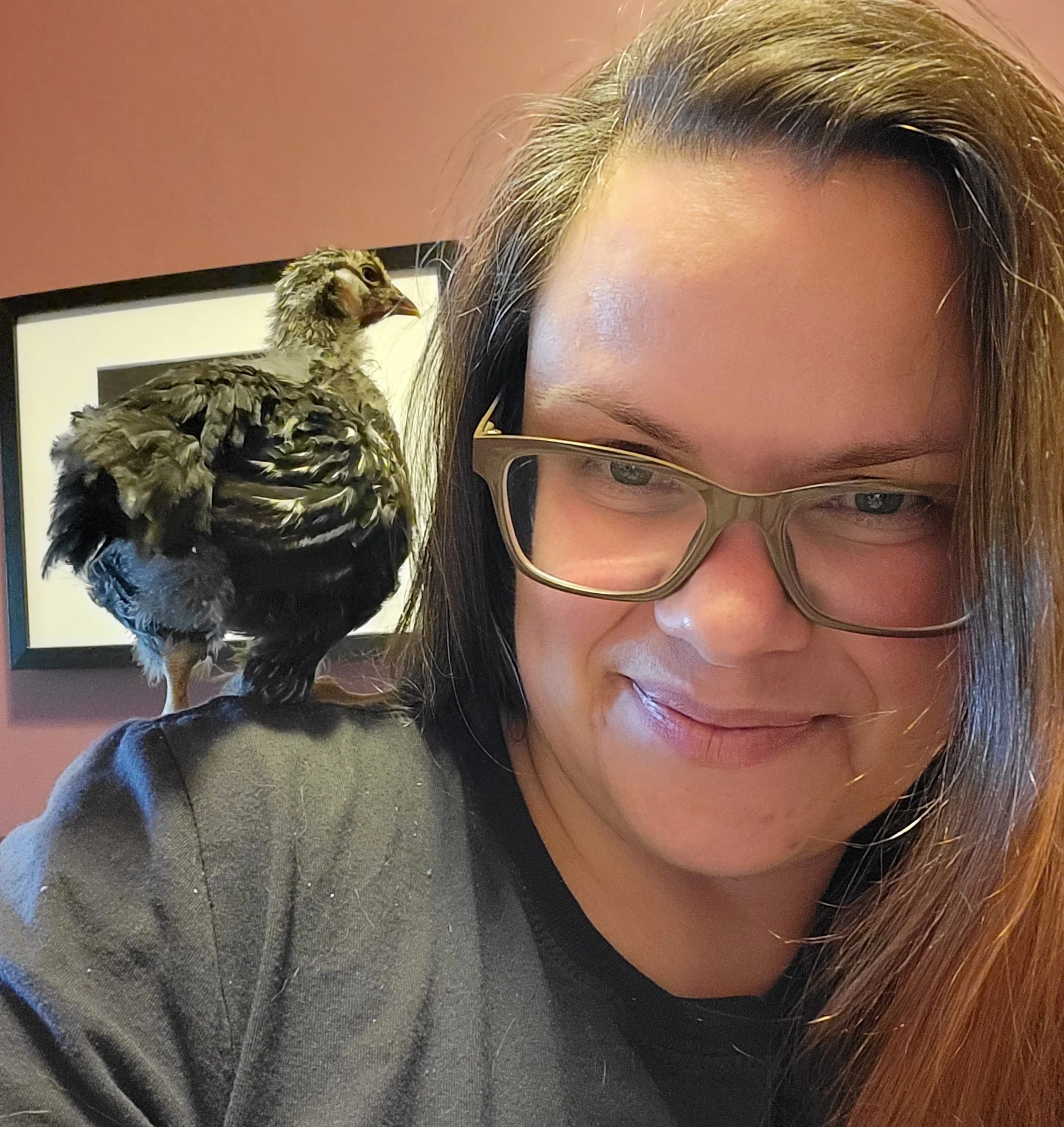 A woman with glasses smiling while a chick perches on her shoulder