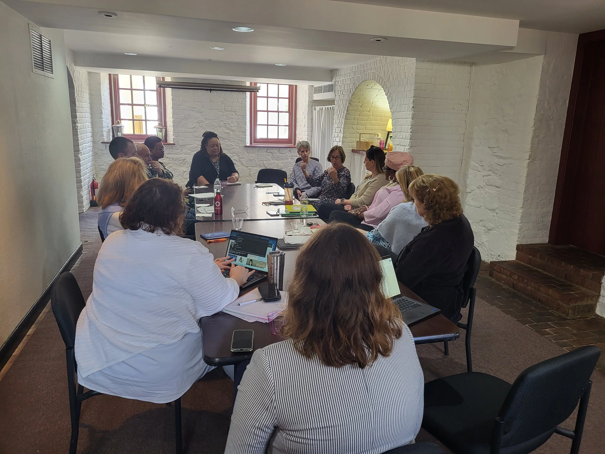 A group of people sitting around a long conference table in a room with white brick walls and windows, engaged in a discussion.