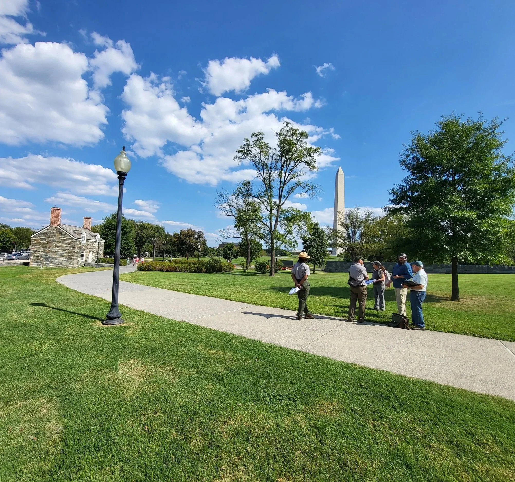Group of people standing on a walkway in a park with trees, grass, and the Washington Monument in the background.