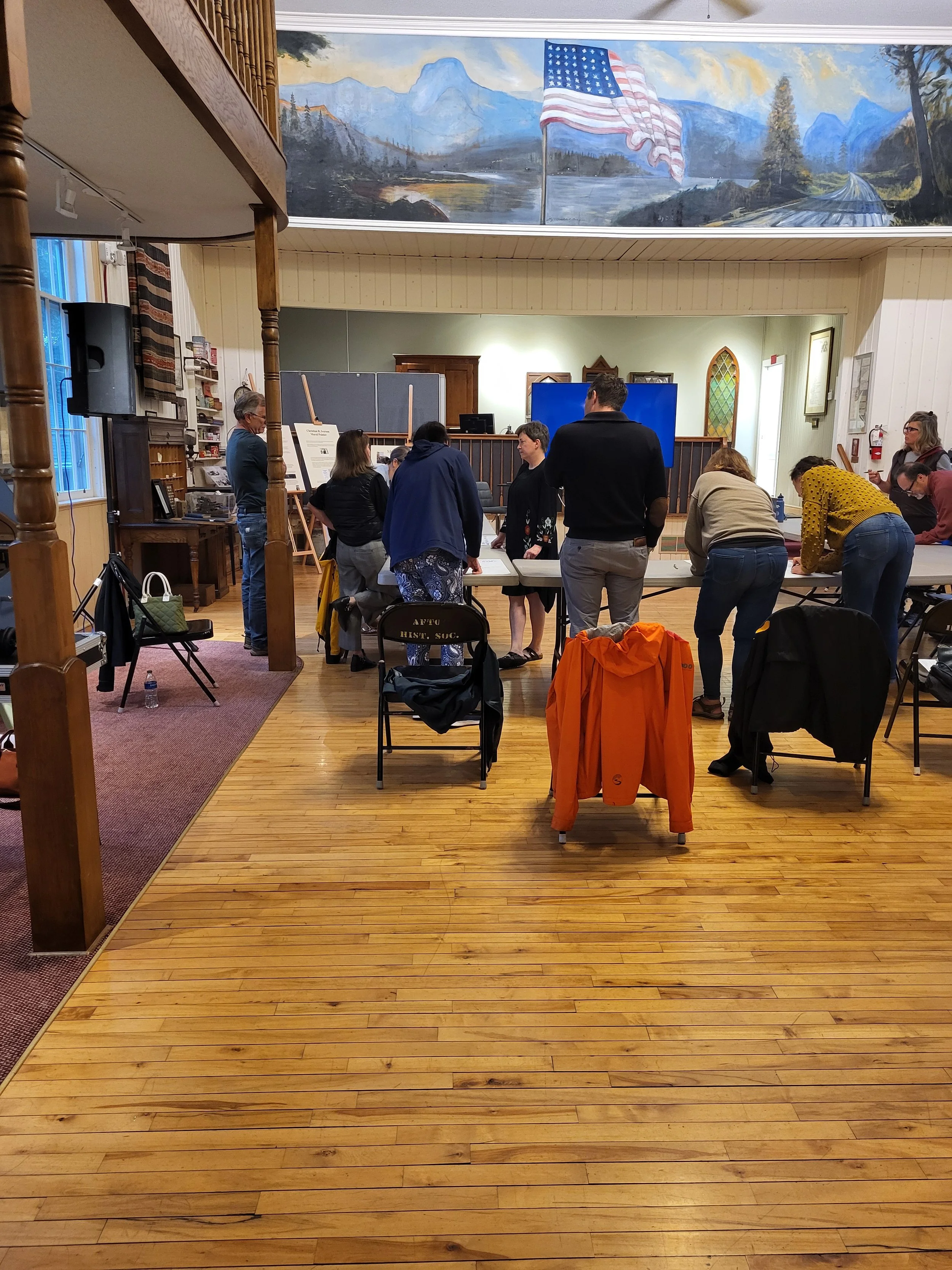 People gathered around tables in a room with a mural of nature and an American flag. There are chairs, a water bottle, and large sheets of paper on the table.