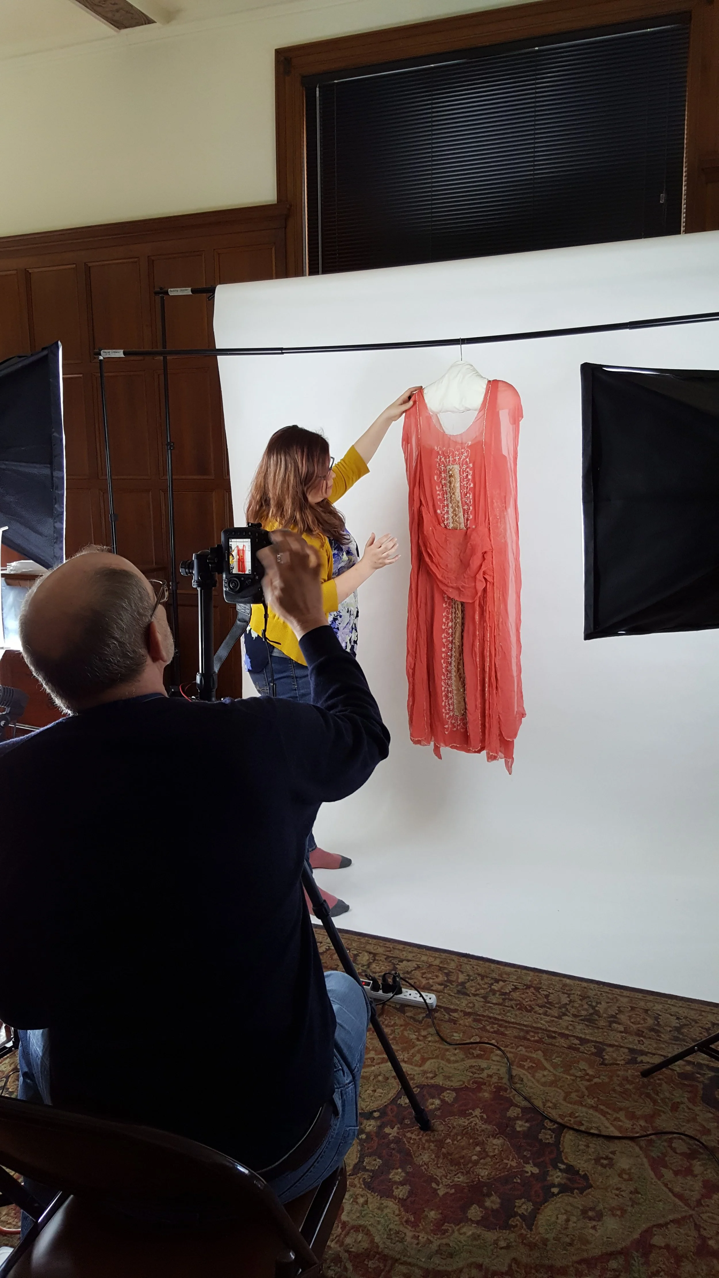 A woman in a yellow cardigan arranging a coral-colored 1920's flapper dress on a hanger in front of a white backdrop. A male photographer focuses a digital camera on a tripod in front.