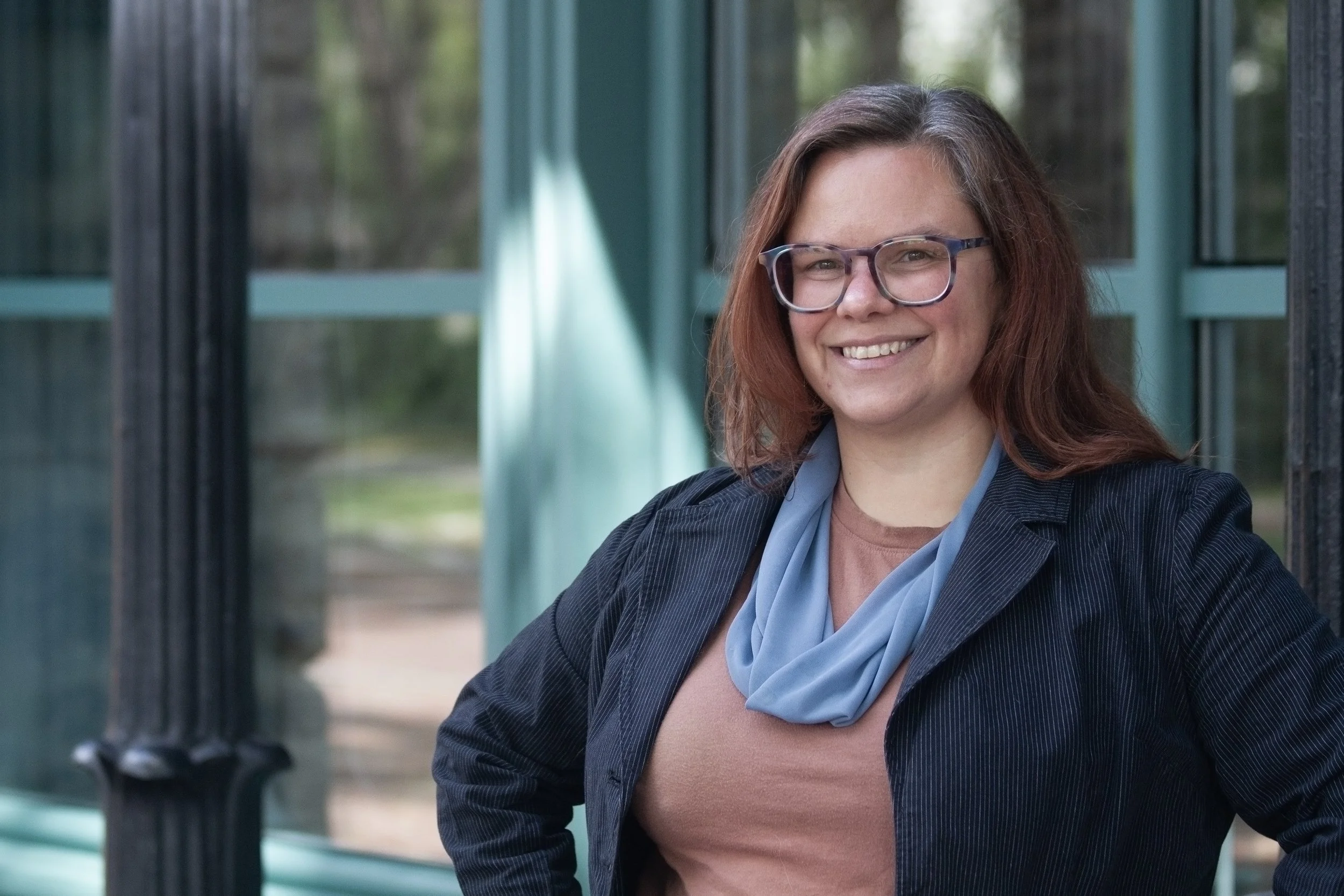 A woman with red hair, glasses, and a blue scarf smiling outdoors, standing near a green and black structure with blurred trees in the background.