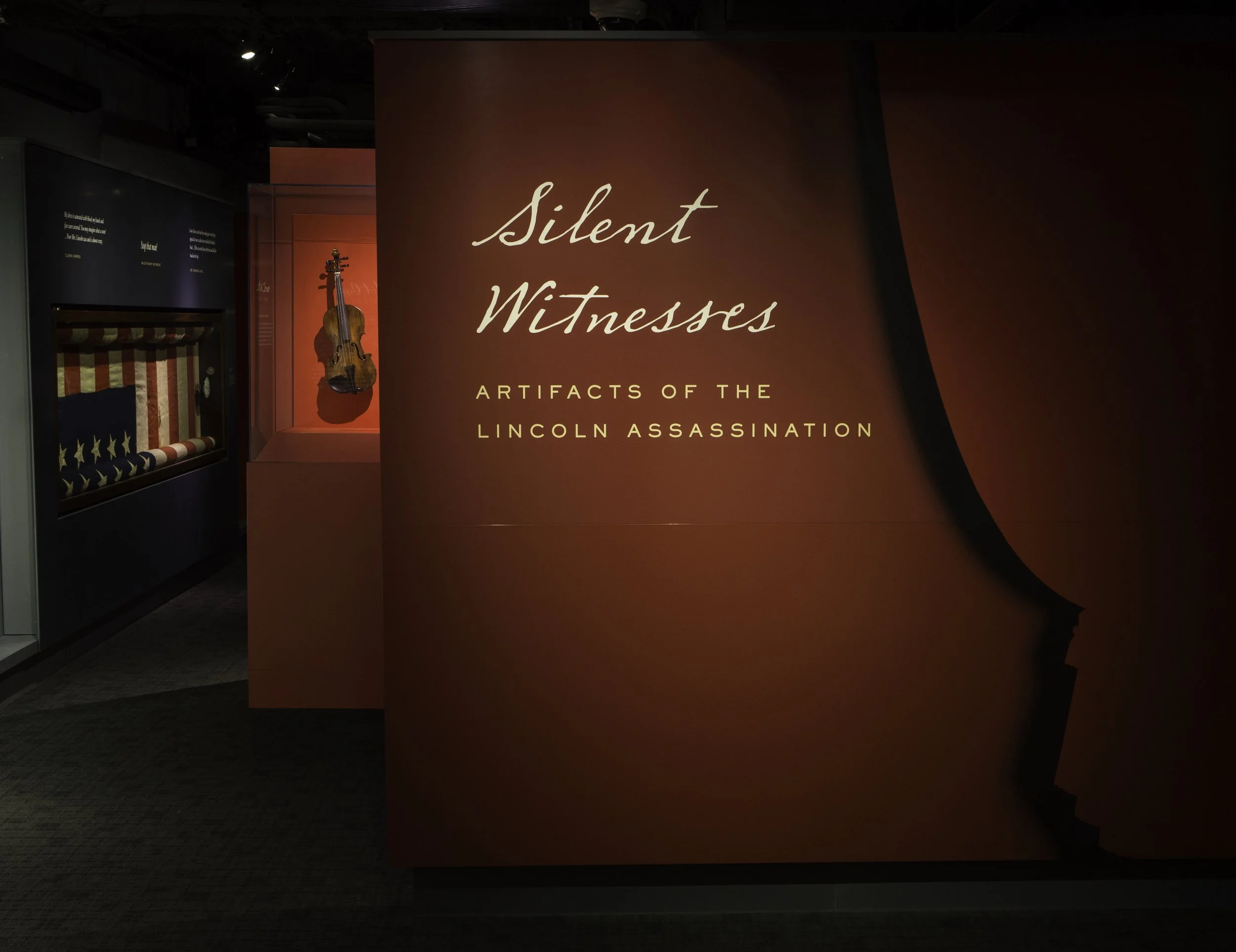 Exhibition display titled 'Silent Witnesses: Artifacts of the Lincoln Assassination' with a red background, a violin, and an American flag coffin in a museum.