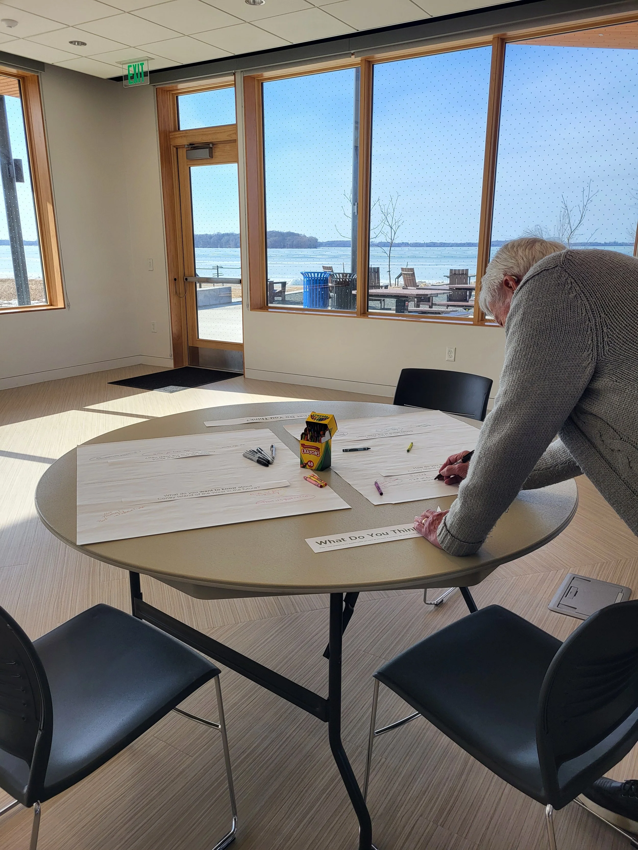 A man in a gray sweater is leaning over a round table with large sheets of paper, markers, and a box of Crayola crayons, in a room with large windows overlooking a lake with outdoor seating.