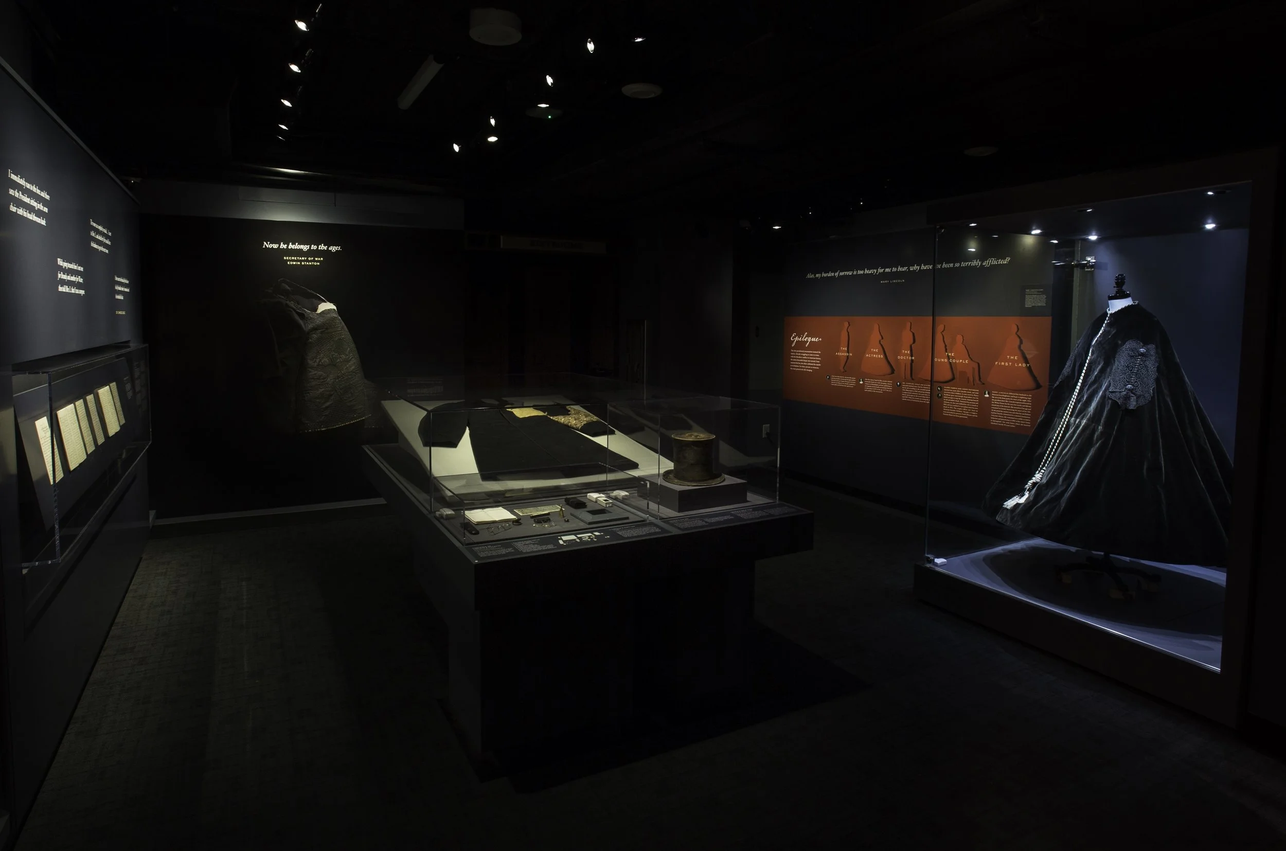Museum exhibit with historical clothing, artifacts, and informational displays in a dark room.