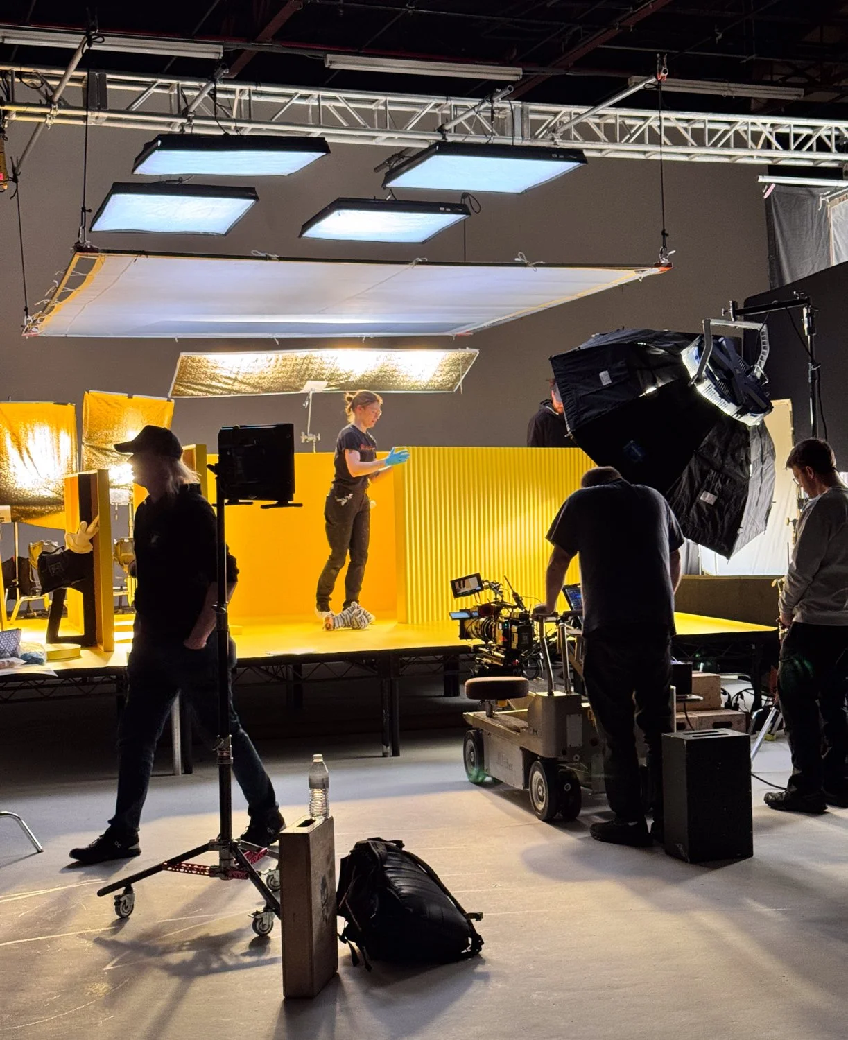 A film or video production set with a yellow backdrop, crew members working around a camera, lighting, and a person standing on the yellow stage. Large soft lights and equipment are overhead, with crew preparing for a shoot.