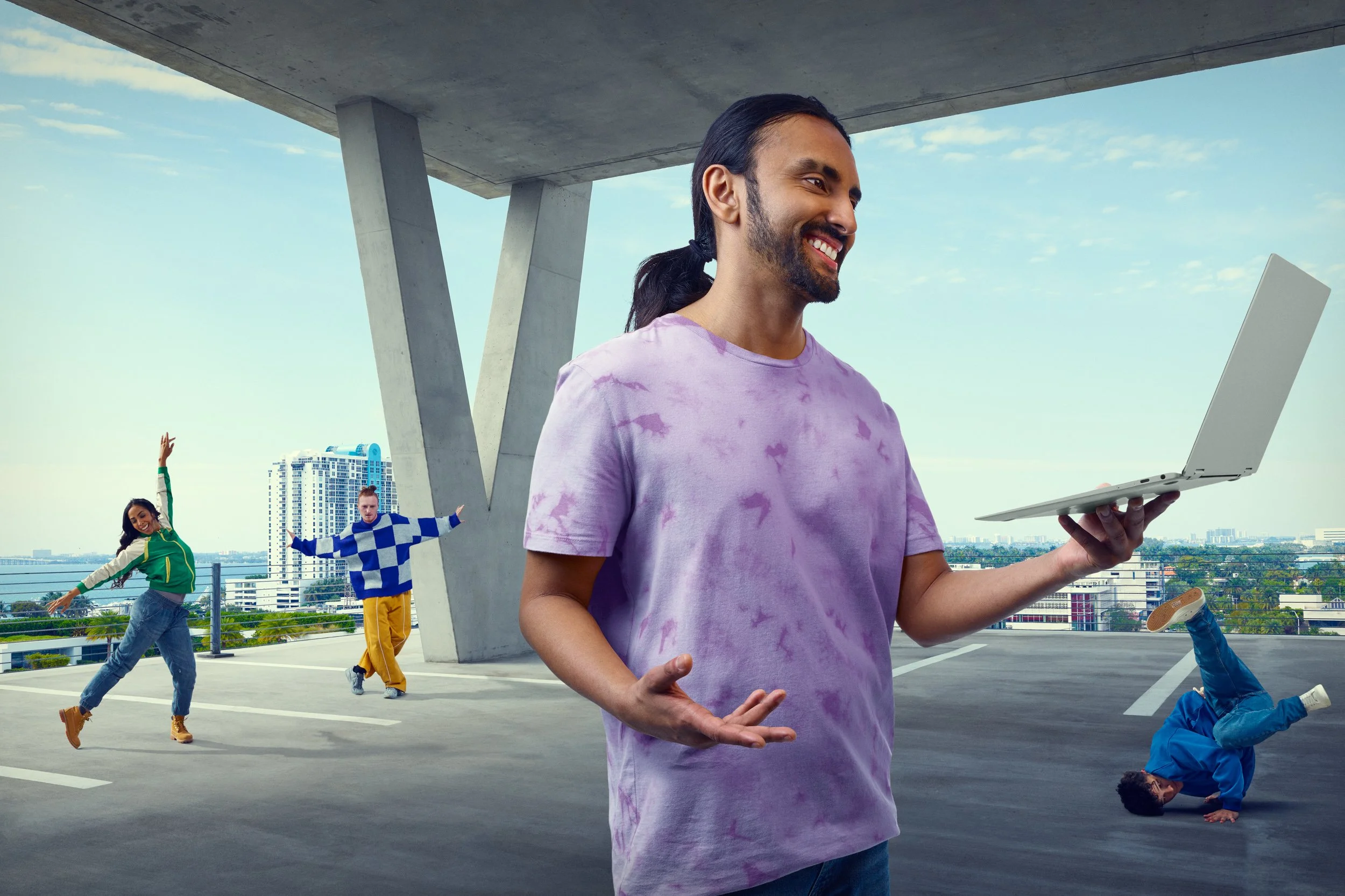 A man with long dark hair in a ponytail, wearing a purple tie-dye T-shirt, smiling and holding a laptop on an outdoor parking garage roof. Behind him, three people are dancing; a woman in a green jacket and jeans, a man in a blue and white checkered 