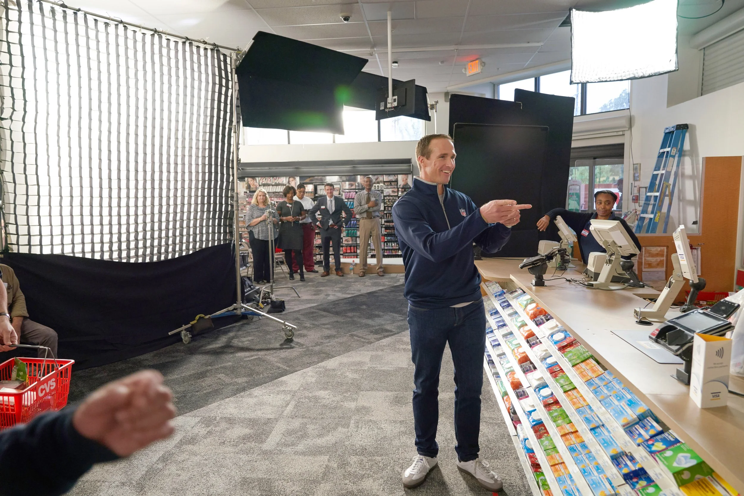 A man in a navy jacket smiling and pointing at the checkout counter in a retail store, with a group of people in the background, and a professional photography setup to the left.