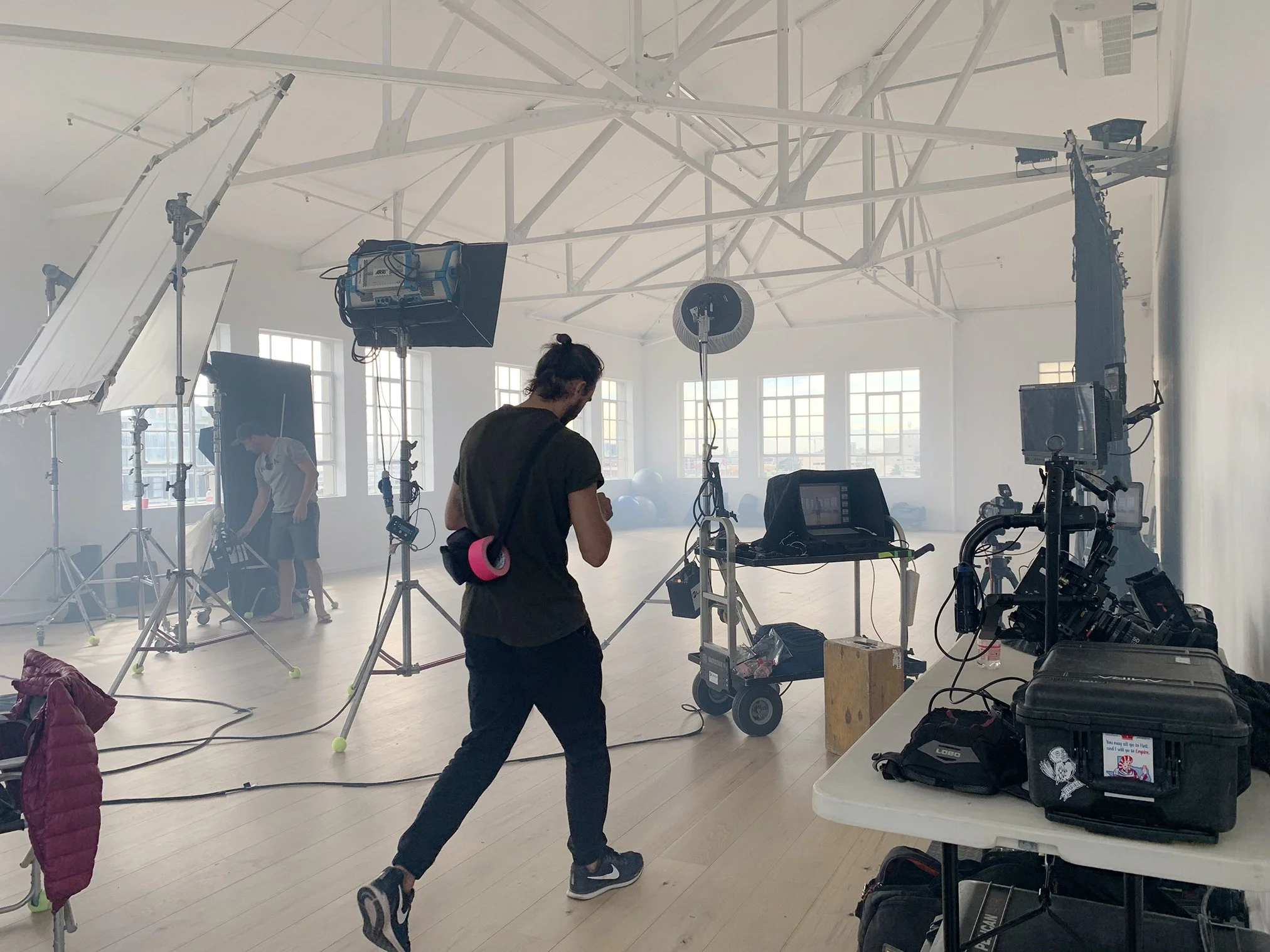 Behind the scenes of a video or photo shoot in a spacious, sunlit studio with large windows. Crew members and equipment such as lights, stands, and cameras are set up around the space. One person is walking through the studio while others are prepari