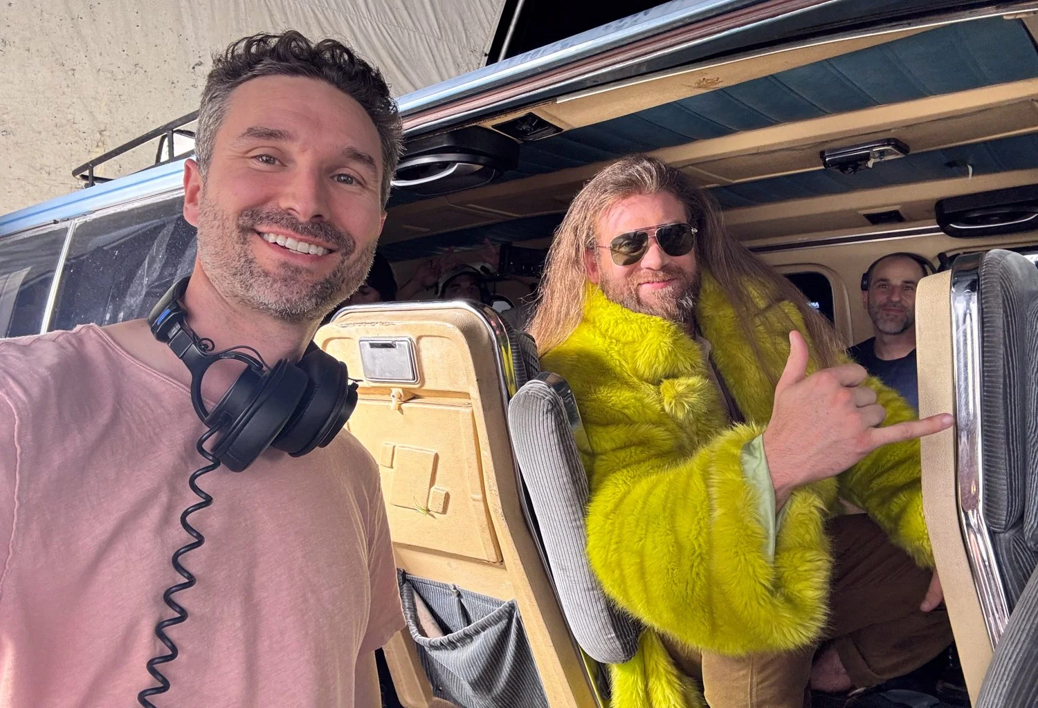 Two smiling men inside an aircraft, with one taking a selfie and the other showing a thumbs-up, surrounded by other people.
