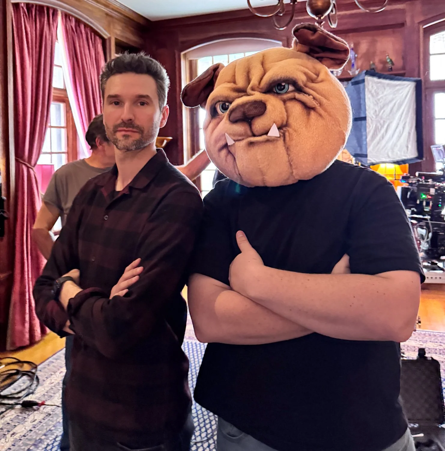 Two men standing indoors with crossed arms. One is wearing a mask of a bulldog with blue eyes and fangs.
