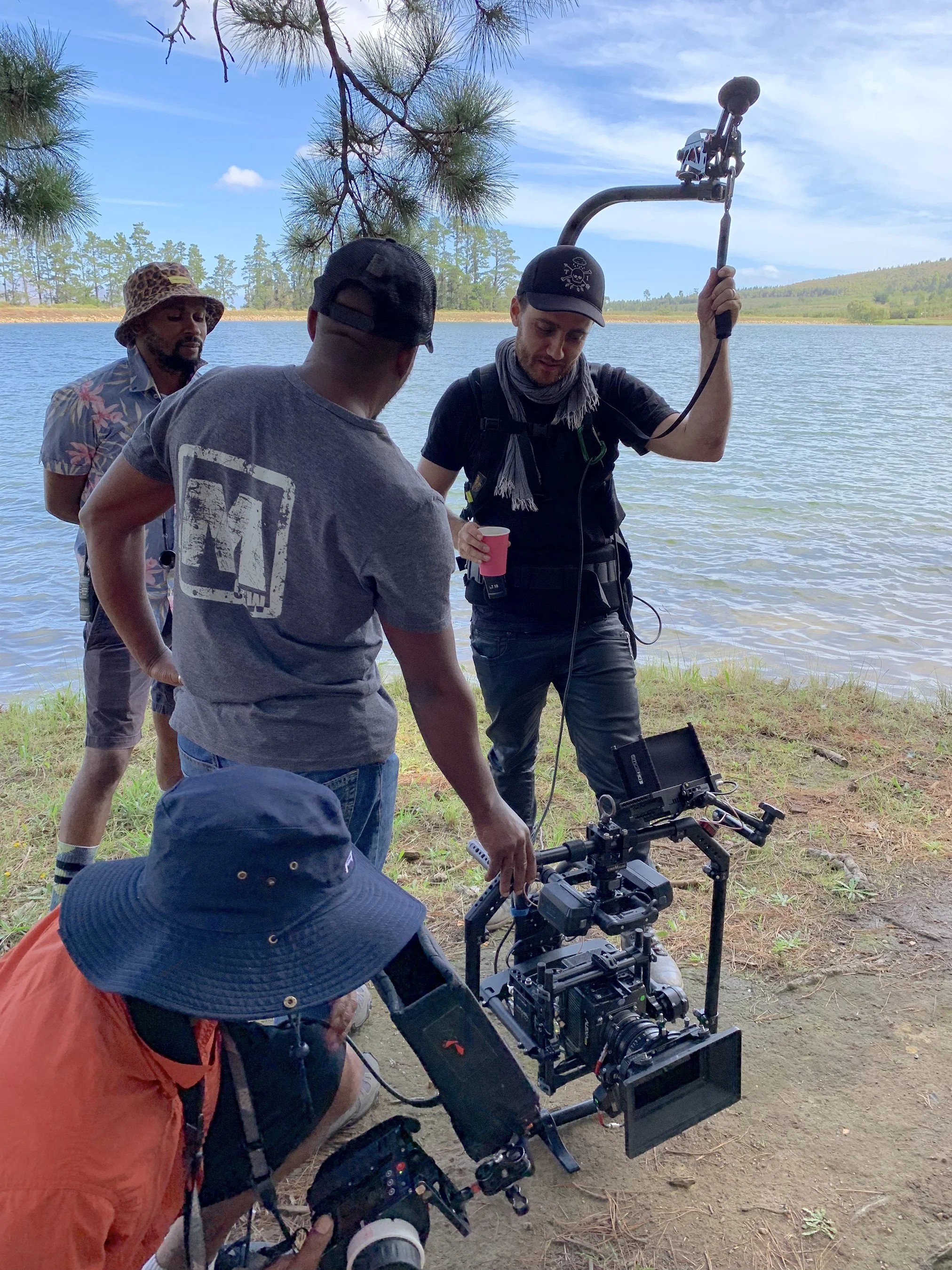 Film crew on location near a lake, with a camera rig and crew members setting up a shot, and one person holding a boom microphone.