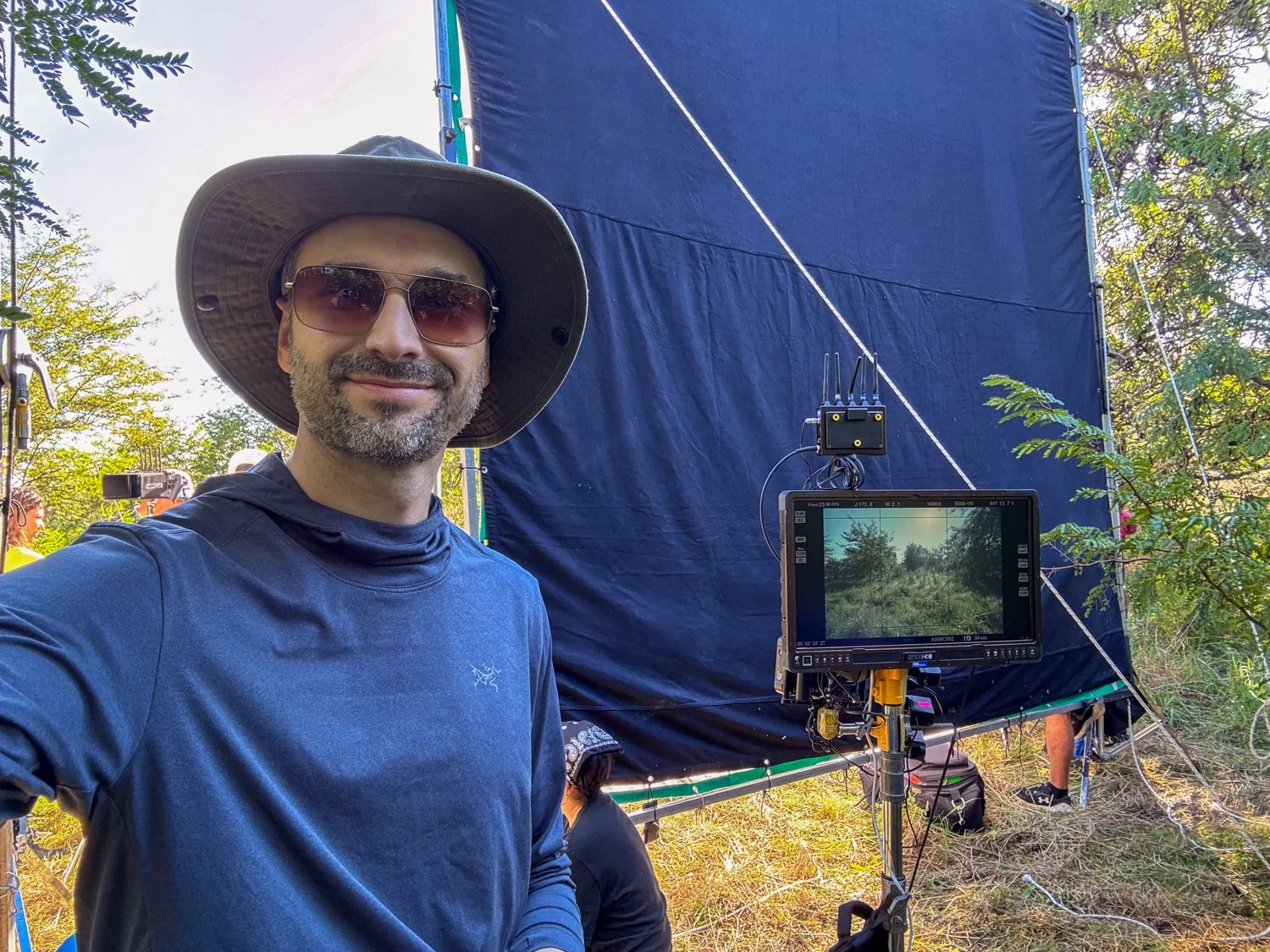 A man wearing sunglasses, a wide-brimmed hat, and a blue long-sleeve shirt taking a selfie in front of a film screen outdoors. There’s a monitor on a stand showing the scene he’s filming, surrounded by trees and greenery.