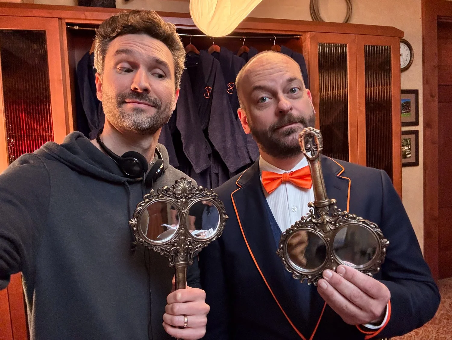 Two men taking a selfie indoors, each holding vintage-style mirror and key objects. The man on the left wears a dark hoodie and headphones around his neck, while the man on the right wears a dark blazer with orange trim, a white shirt, and an orange