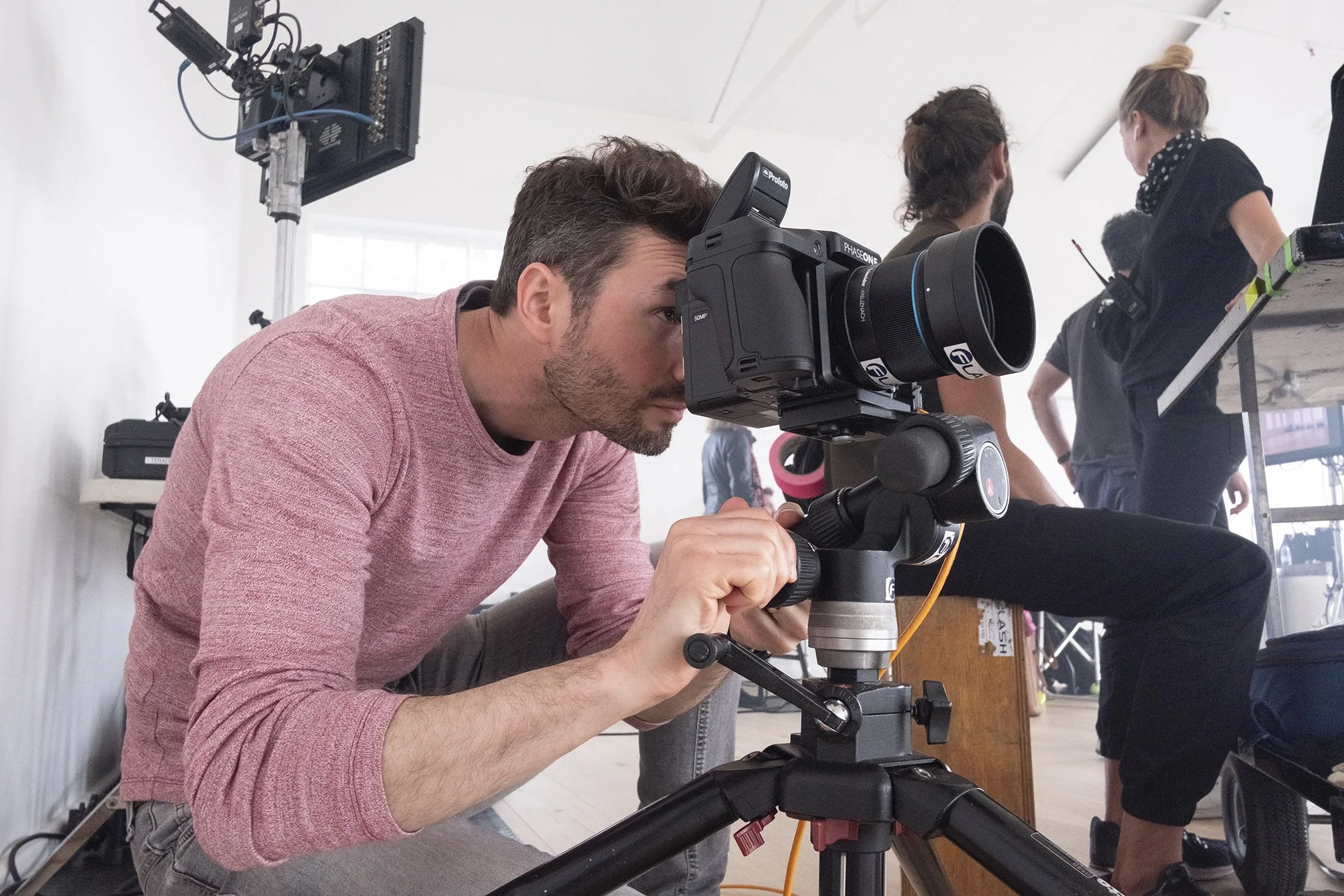 A man operating a professional camera mounted on a tripod during a photoshoot or filming session, with a few people working or talking in the background inside a bright studio.