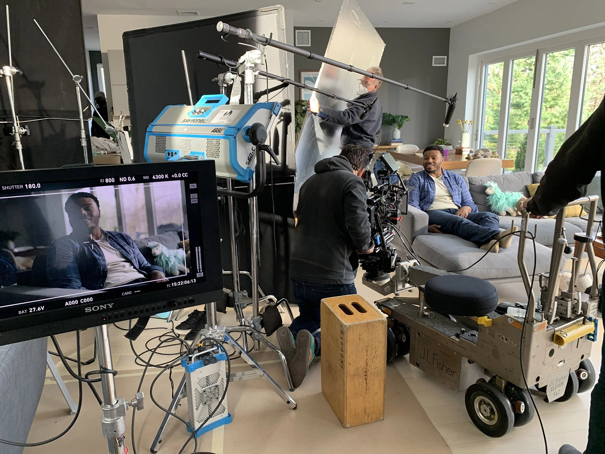 Behind the scenes of a film or TV set showing a man sitting on a gray couch during an interview, crew members operating camera and lighting equipment in a well-lit living room with large windows.