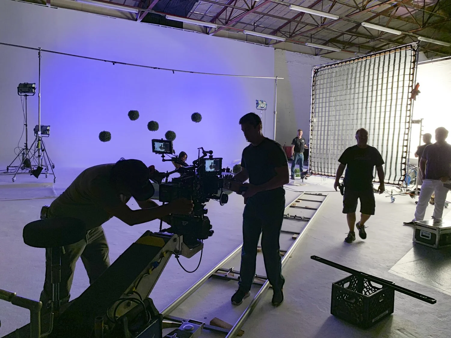 Behind the scenes of a video or photo shoot with crew preparing equipment in a studio with a white backdrop and lighting.