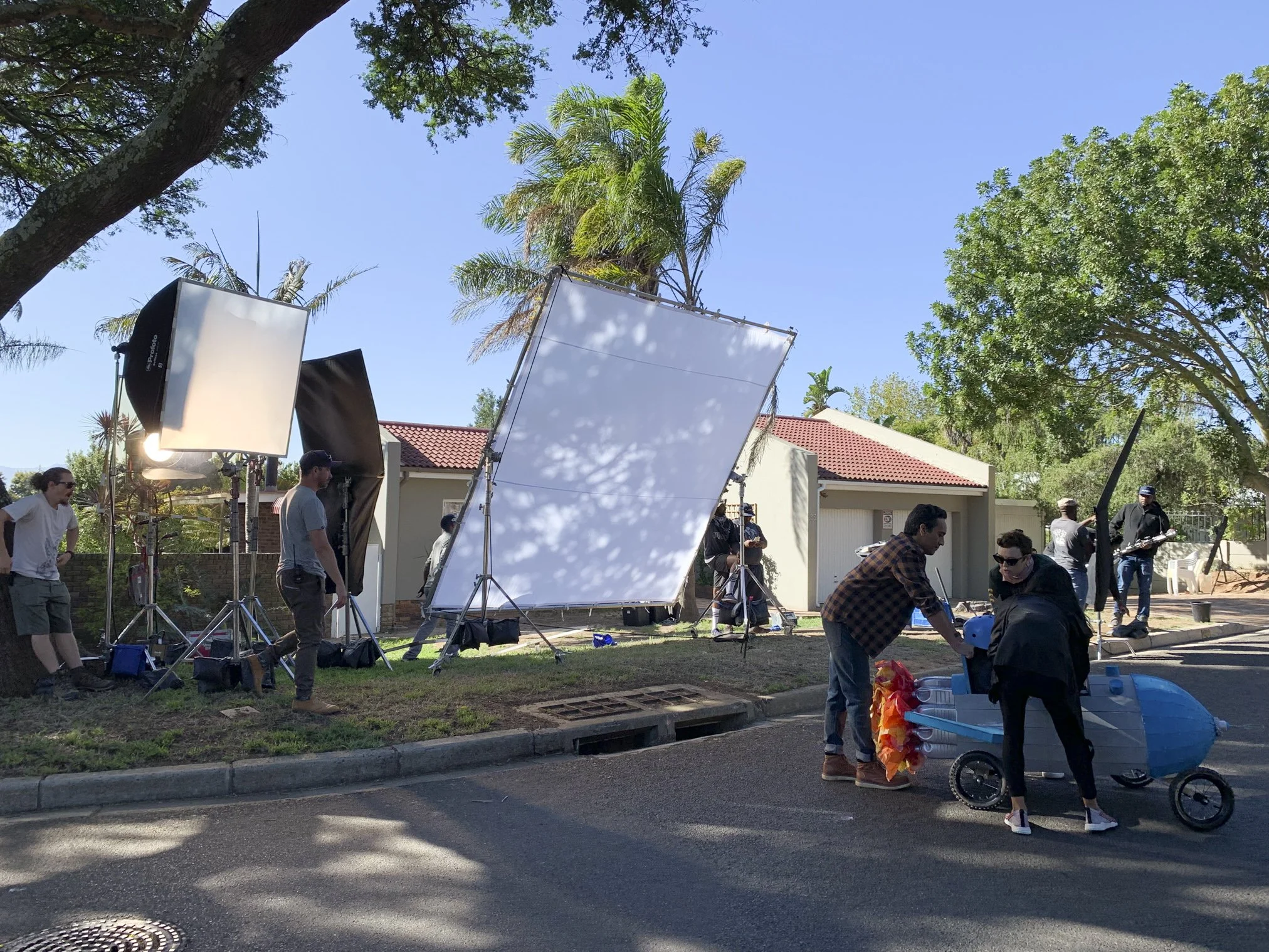 Filming crew setting up outdoor scene with lighting and reflectors, two people with a pool float in a small pool, and crew members in casual clothing, on a residential street with trees and houses, during daytime.