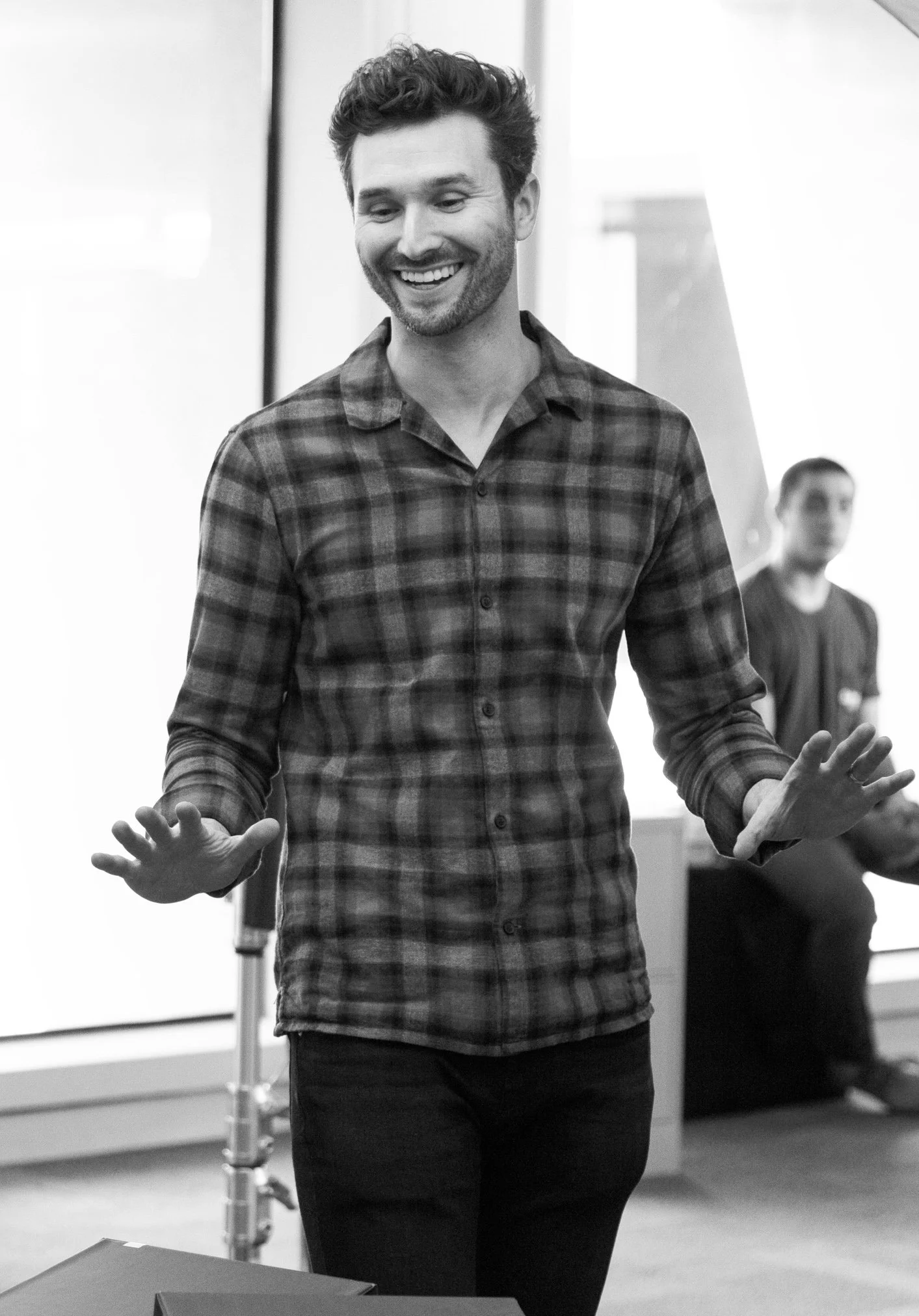 A man smiling and gesturing with his hands in a professional setting, with a window and another person in the background.