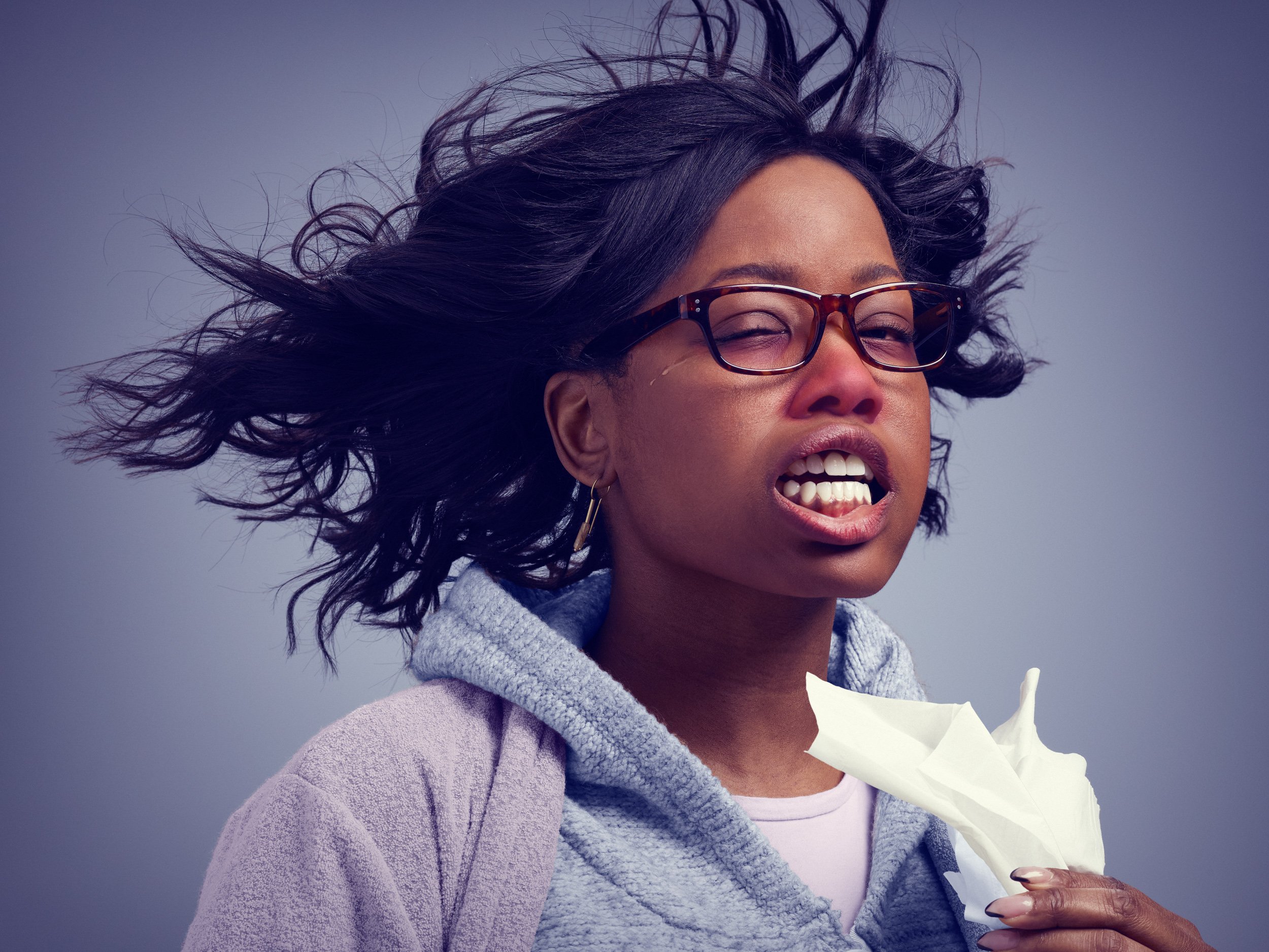 A woman with wavy hair blowing in the wind, wearing glasses, holding a tissue to her mouth, with a distressed facial expression.