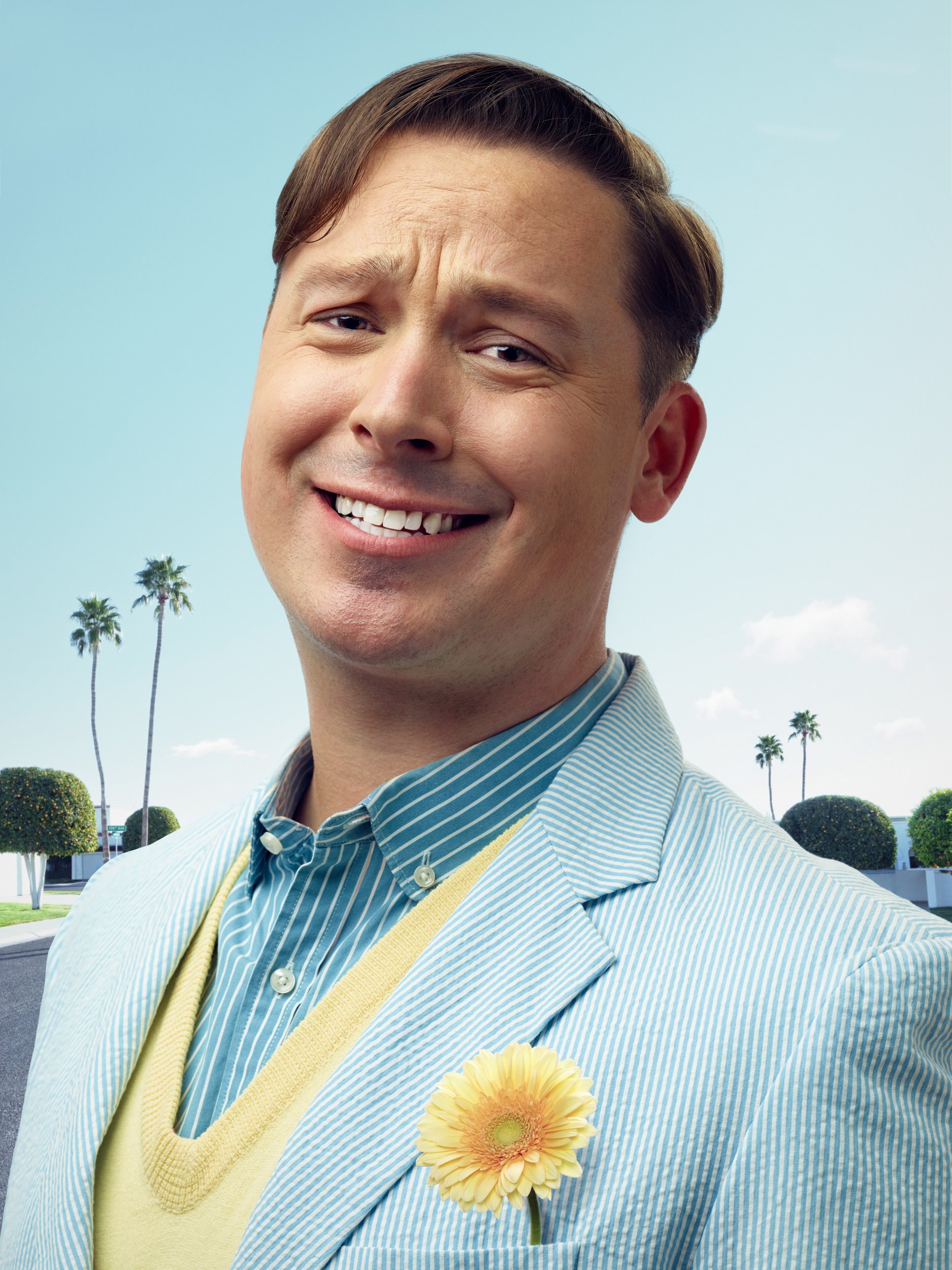 A man smiling outdoors with palm trees in the background, wearing a light blue blazer and a striped shirt, with a yellow flower pinned to his blazer.