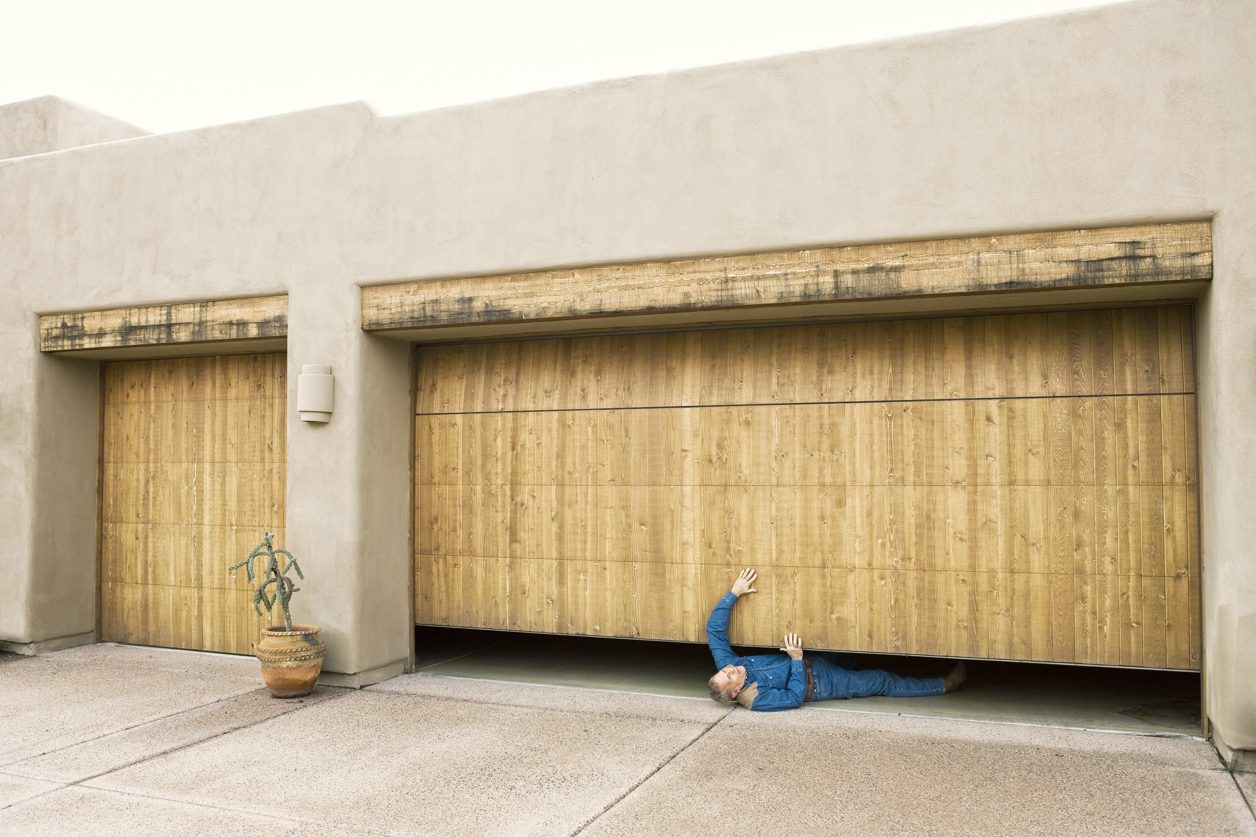 A man lying on the ground under a closed wooden garage door, holding the edge of the door, with a potted cactus near the garage wall.