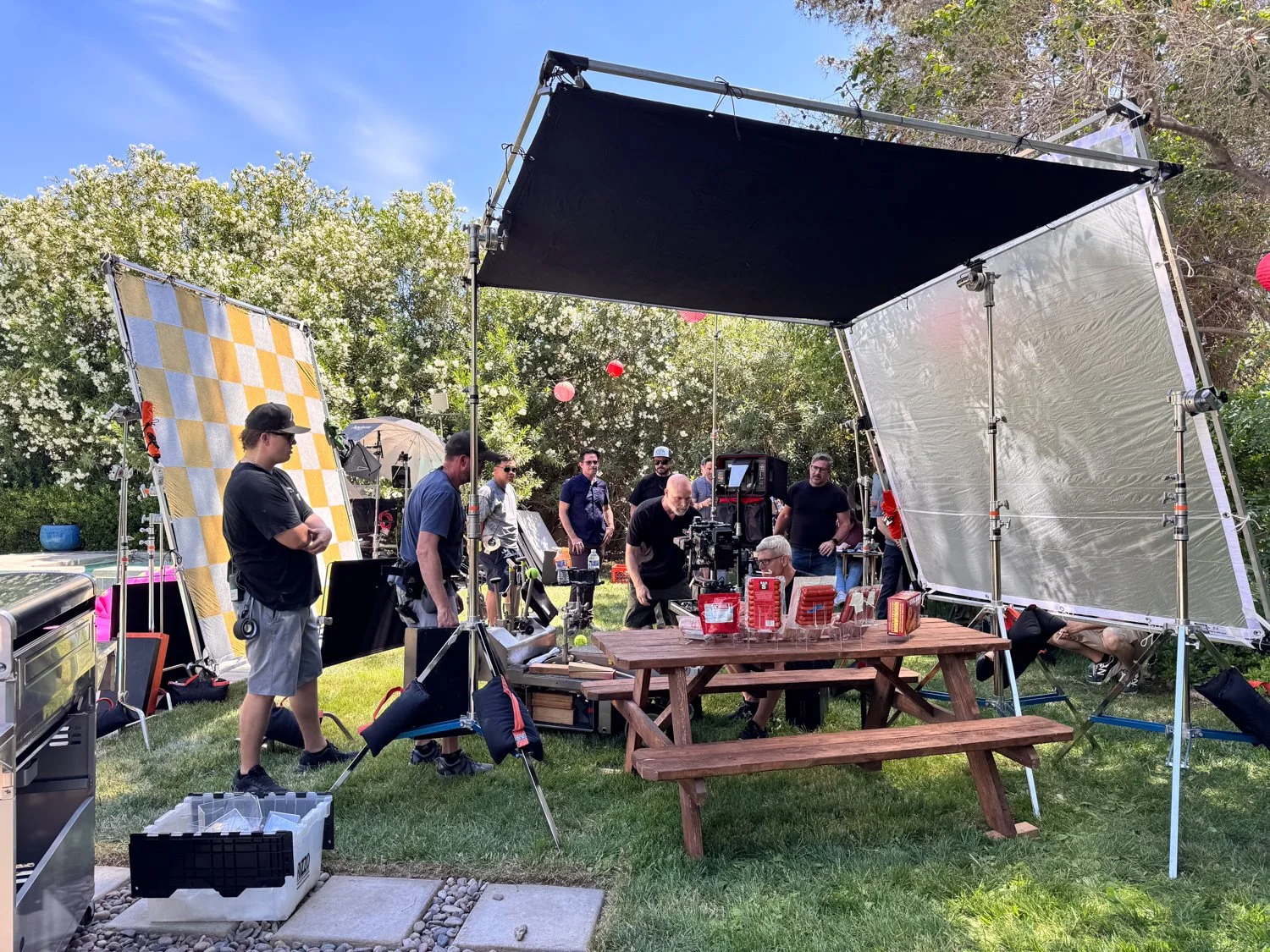 Filming crew setting up equipment outdoors on a sunny day with trees, a picnic table, and various filming equipment including large lighting and reflector panels.