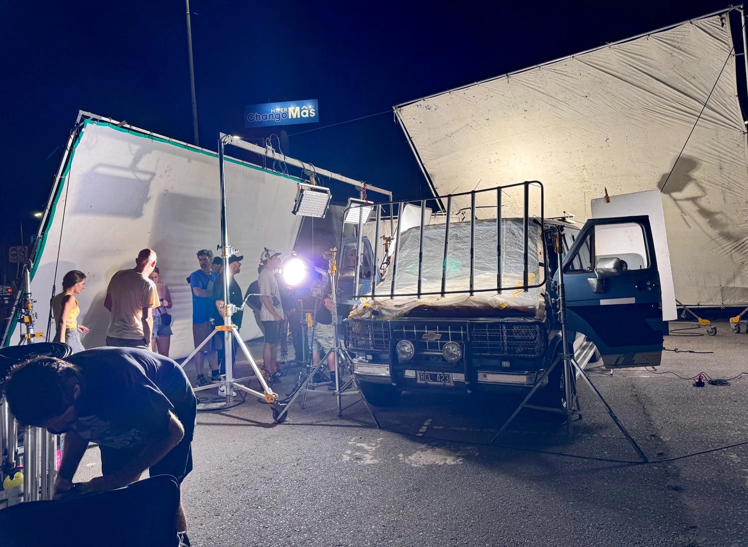 Film crew filming a scene on a street at night with professional lighting, a truck covered with a white sheet, and large reflectors.