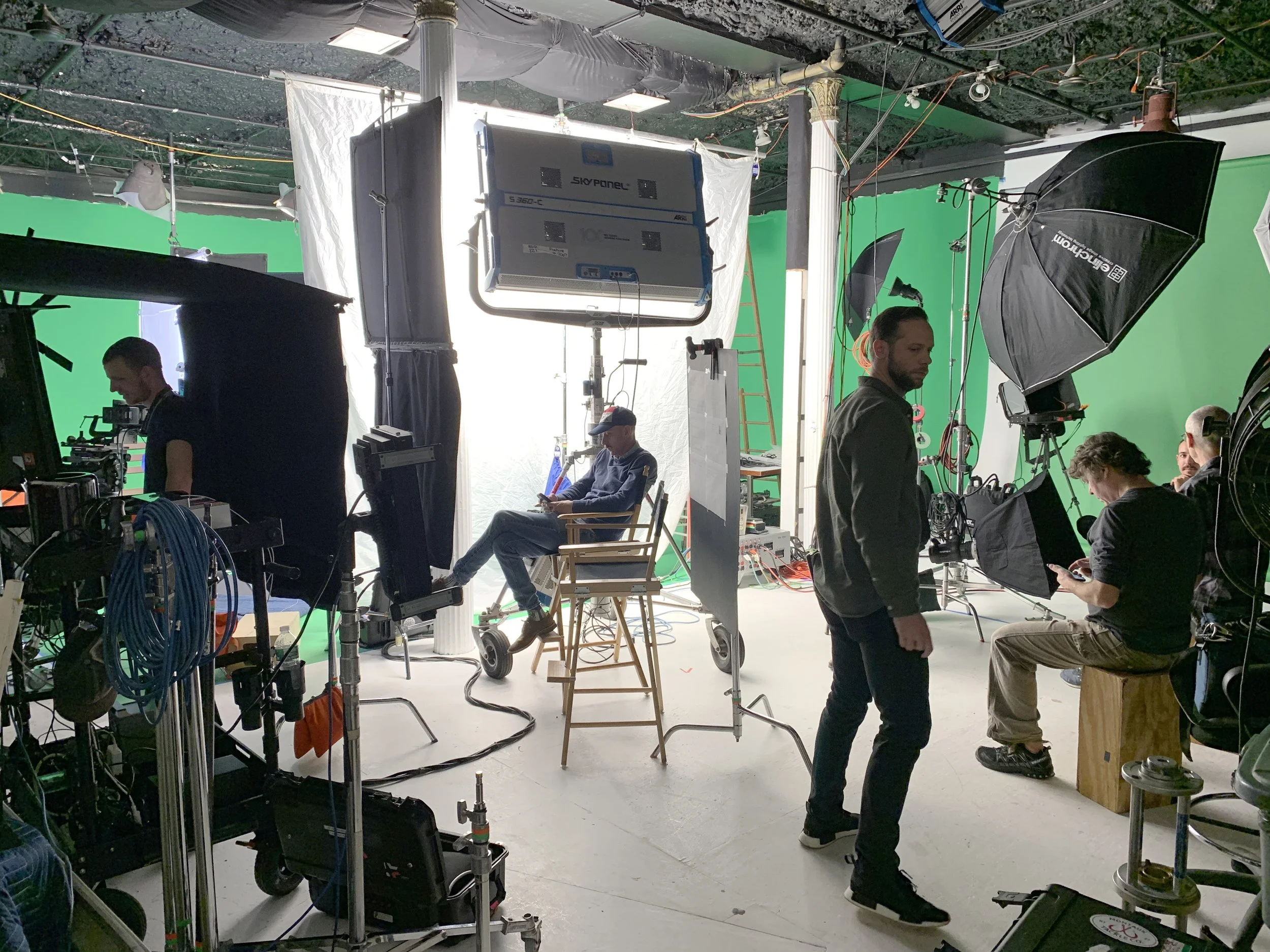 A film or video production set with a green screen background, crew members working with equipment, lighting, and monitors.