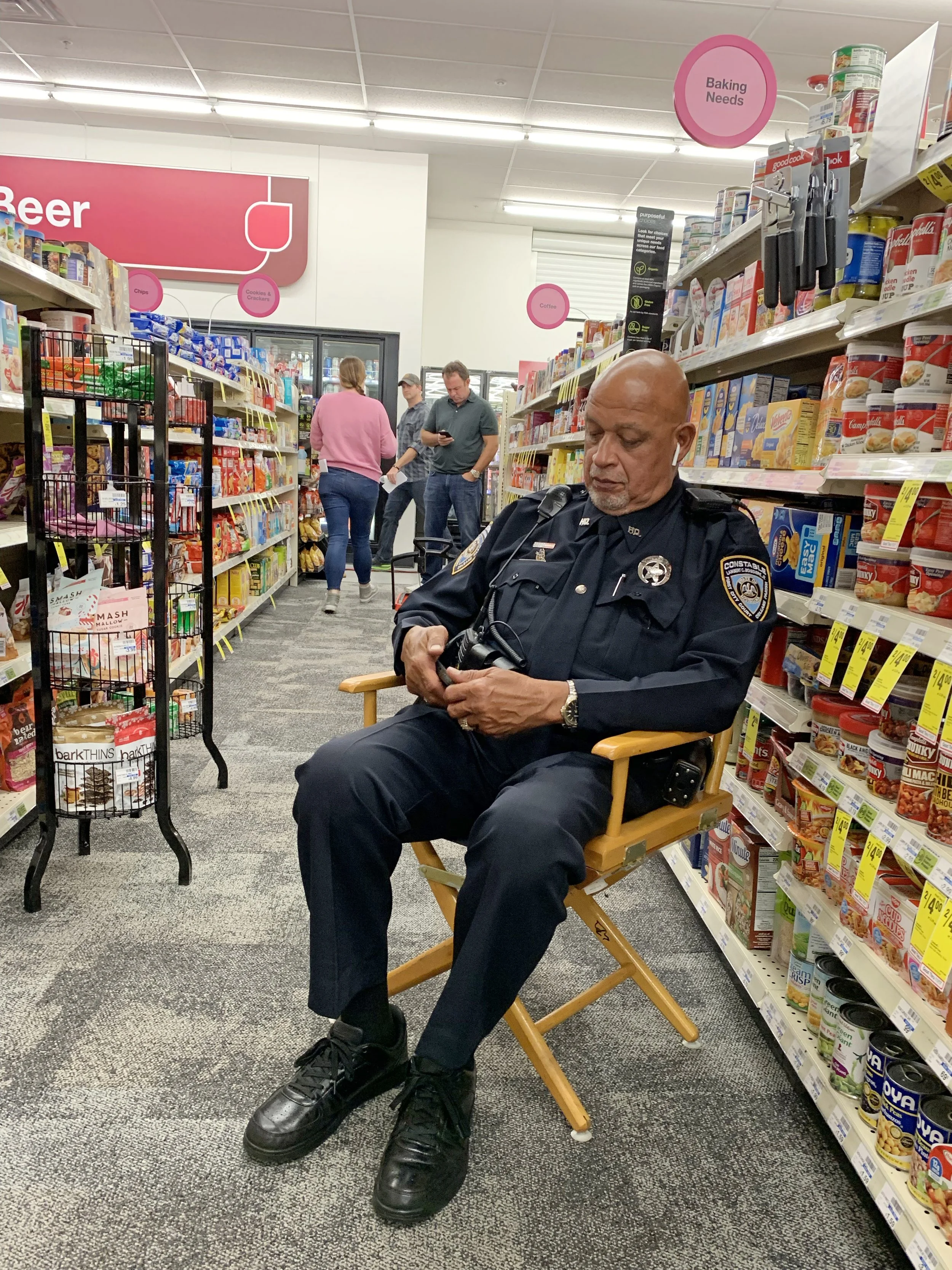 A police officer sitting on a folding chair inside a grocery store aisle, looking at his phone. The store shelves are stocked with various food products, and other shoppers are in the background, with signs indicating sections like 'Baking Needs' and