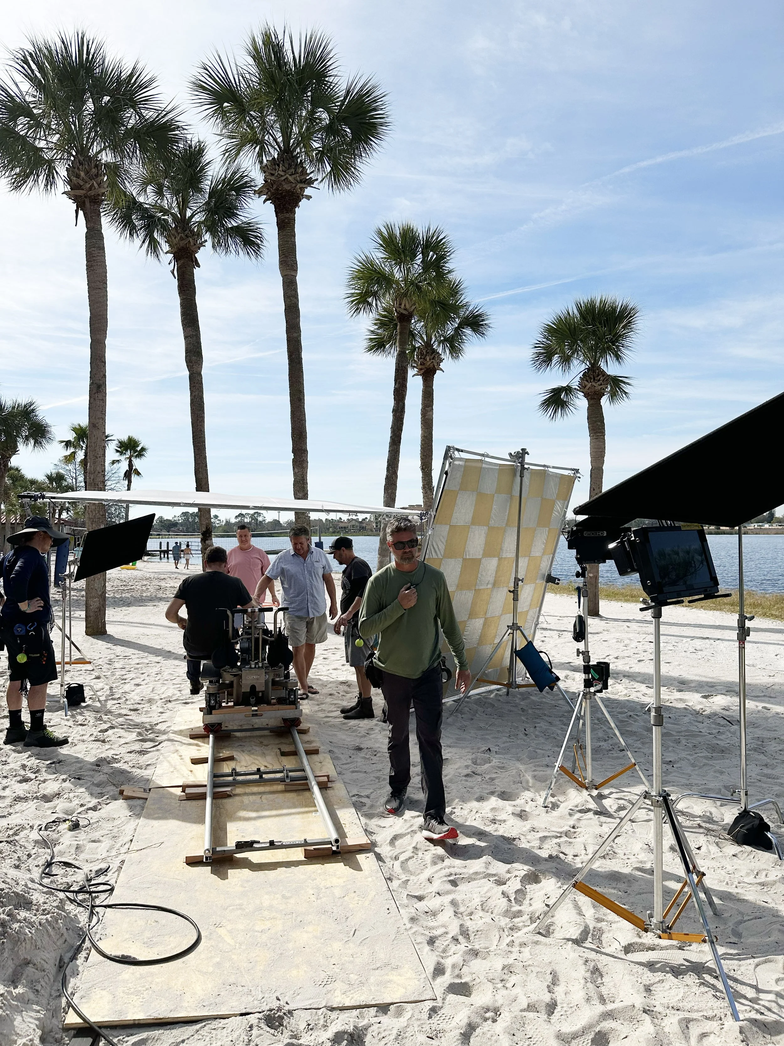 Filming crew on a beach setting up filming equipment with palm trees in the background.