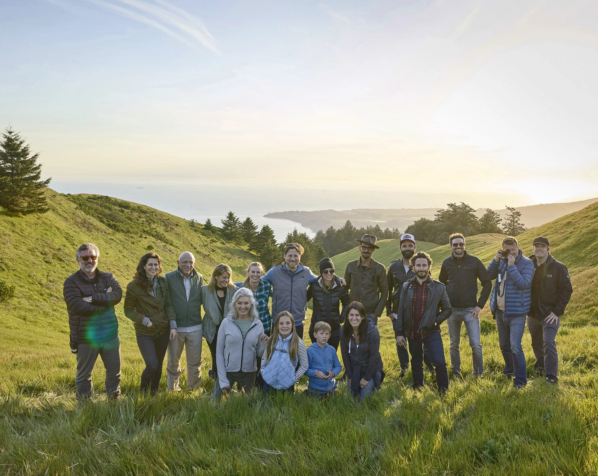 Group of people standing on grassy hill with scenic landscape of trees, distant water, and sunset sky.
