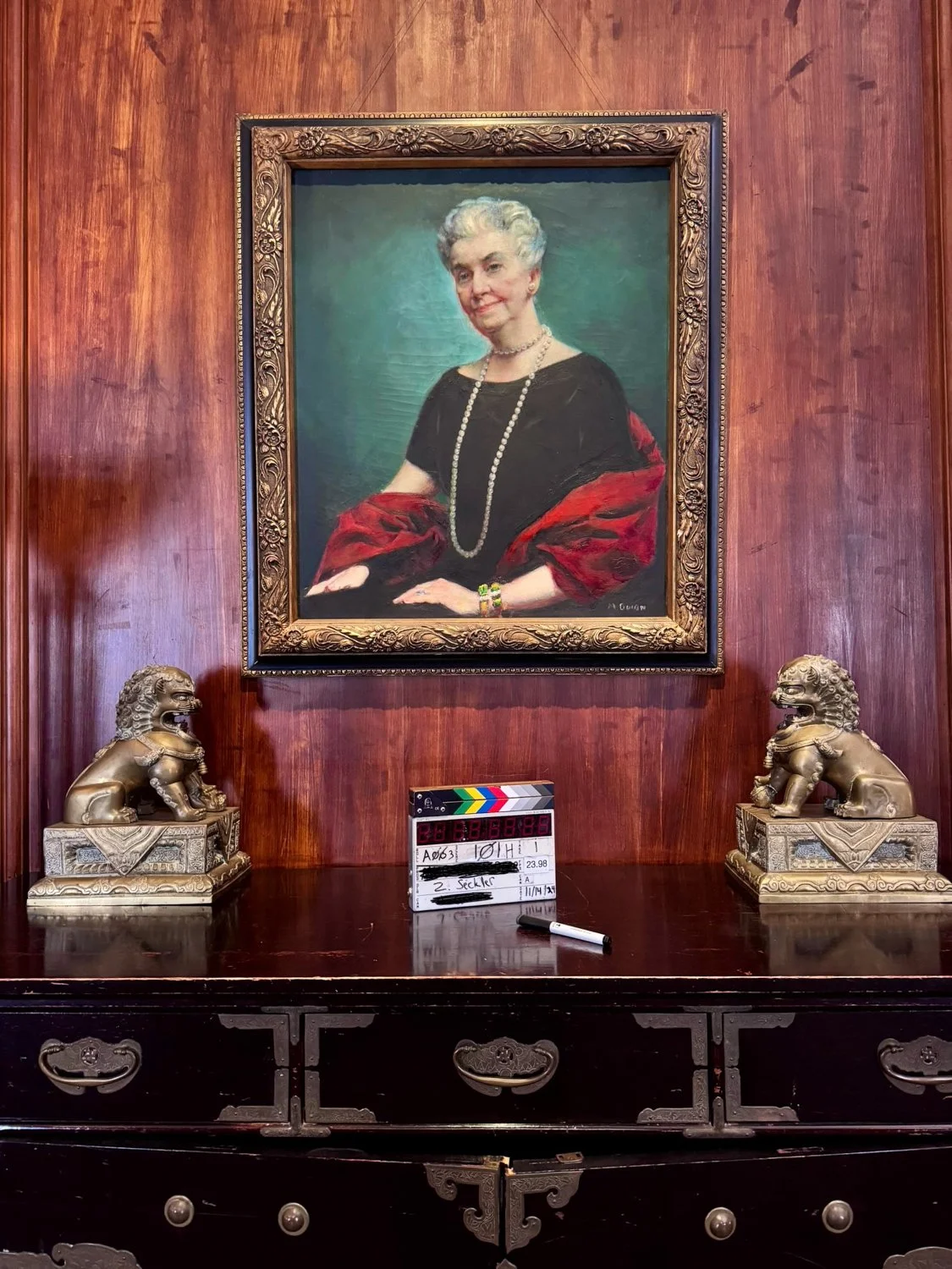 A framed portrait of an elderly woman with short, gray hair wearing a black dress with a rosé-colored shawl, multiple necklaces, and bracelets, hanging on a wood-paneled wall. Below the portrait is a dark wooden dresser with two ornate lion statues,