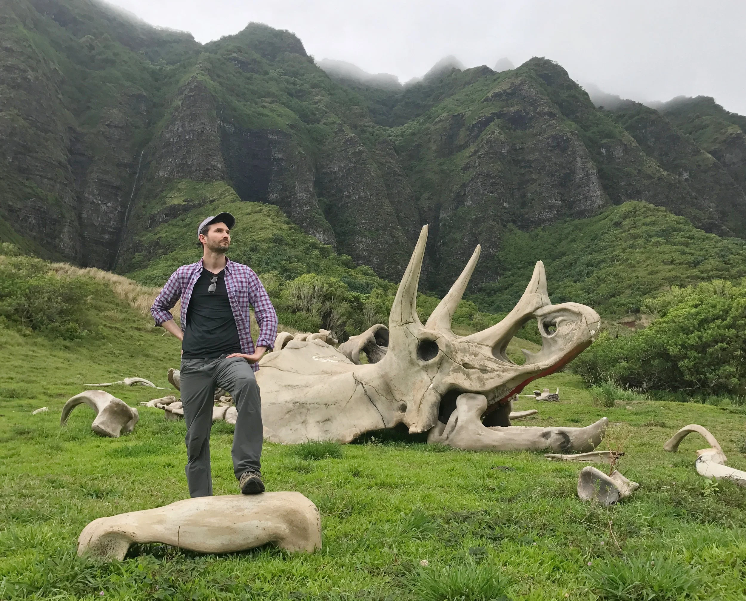 A man standing outdoors in front of a large dinosaur skull fossil with mountainous greenery and misty clouds in the background.