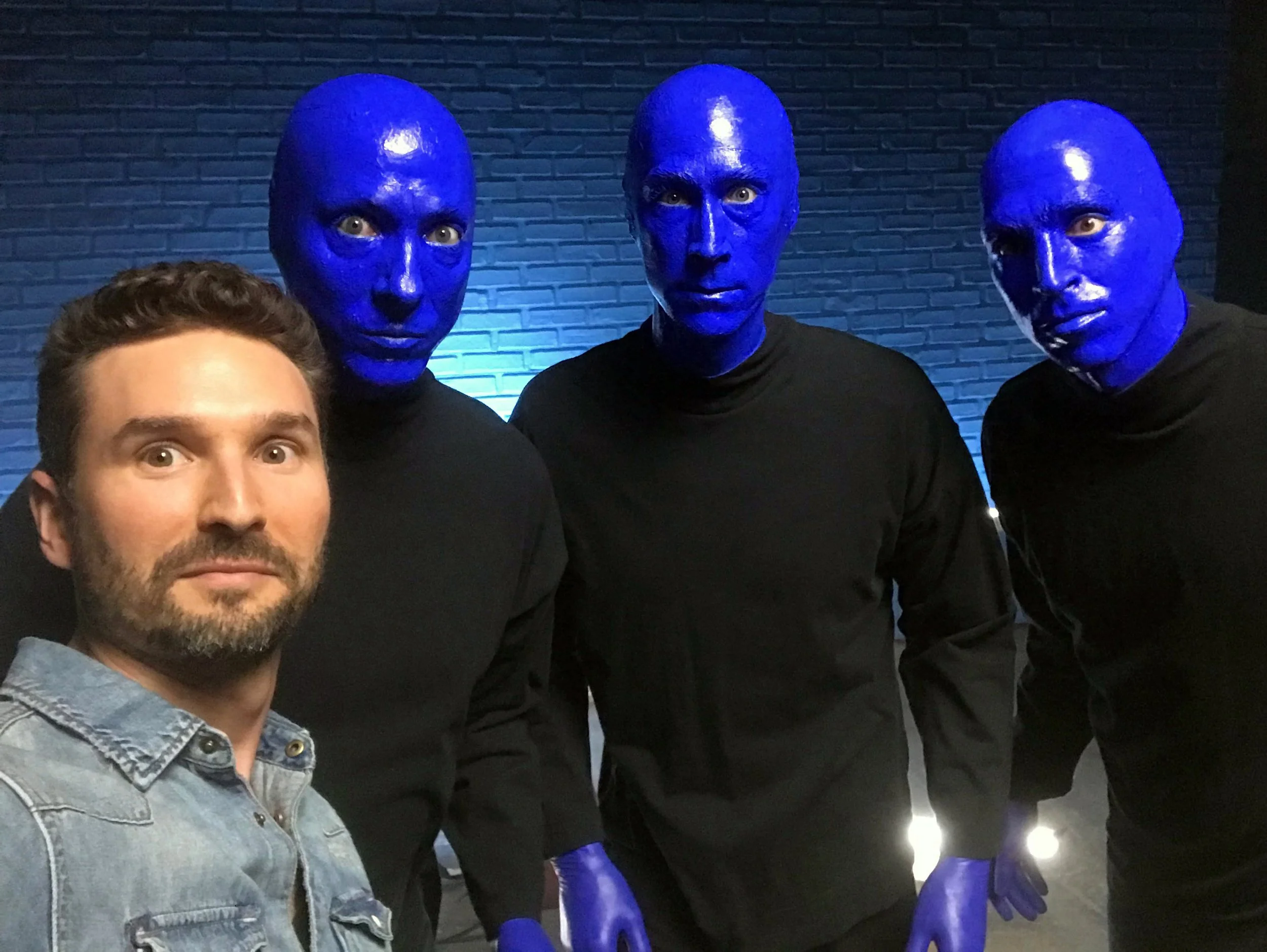A man taking a selfie with three people dressed as blue-skinned aliens with bald heads and black clothing, standing against a dark brick wall.